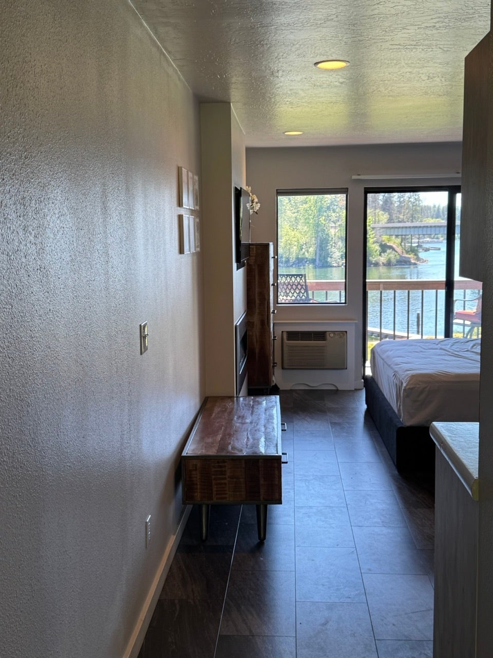 Interior living space with dark tile flooring, a window providing natural light, a sliding glass door leading to a balcony, and a scenic water backdrop featuring a bridge