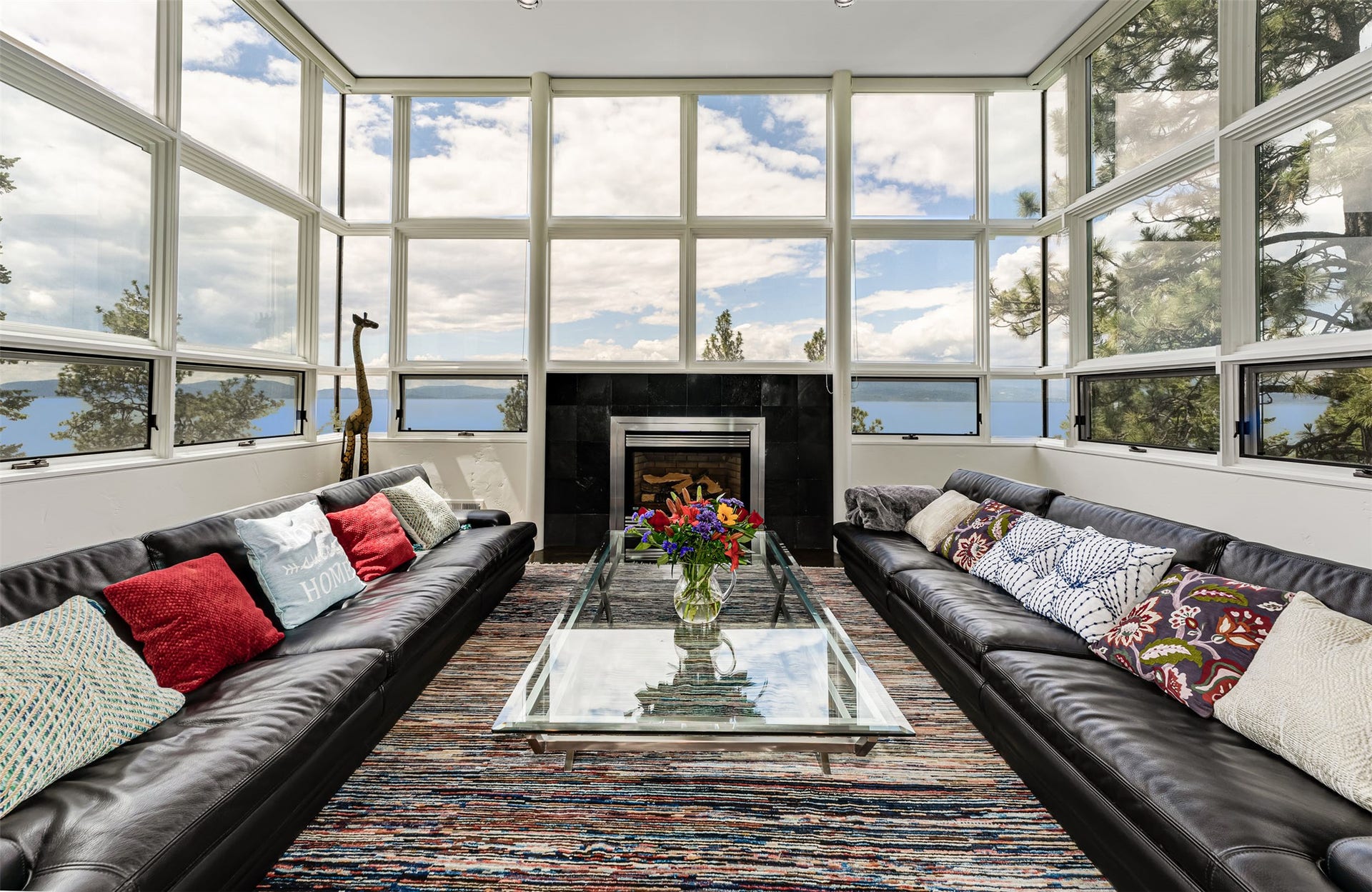 Spacious living area featuring expansive floor-to-ceiling windows, a central fireplace with a dark stone surround. Exquisite sky, lake and mountain views envelop the viewer.
