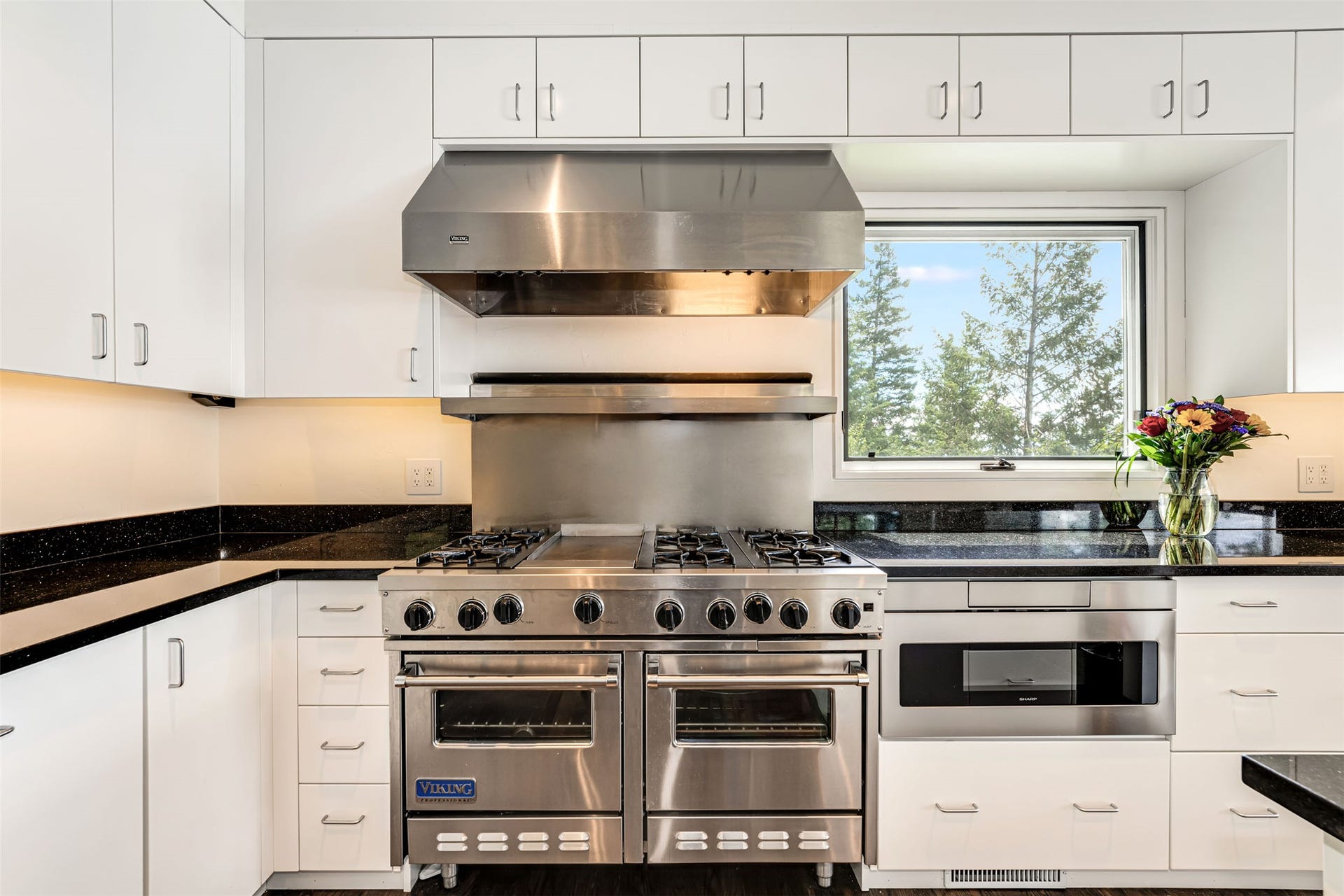 Modern kitchen featuring white cabinetry, bronze flecked granite countertops, and stainless steel appliances, including a professional-grade Viking range with a pair of ovens, 6 gas burners. Next to it is and a built-in microwave drawer.