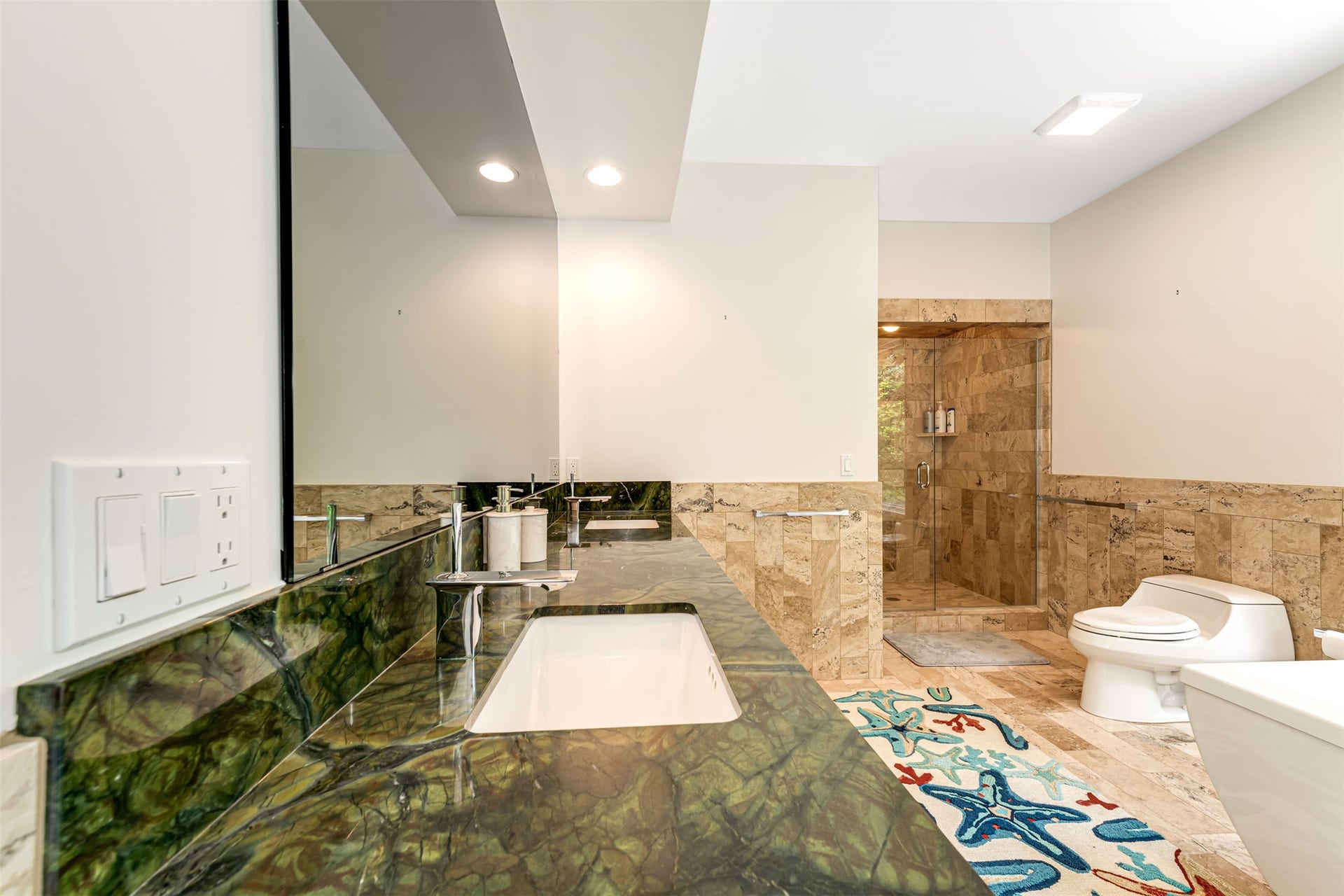 Bathroom featuring a green stone countertop with an integrated sink, a large wall-mounted mirror, recessed lighting, a glass-enclosed shower with tiled walls, and a toilet