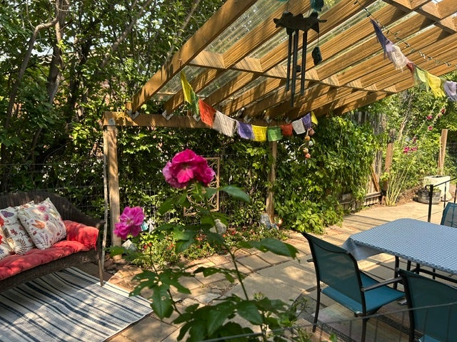 Outdoor patio featuring a wood-frame pergola with a clear panel roof, stone paver flooring, and lush green landscaping