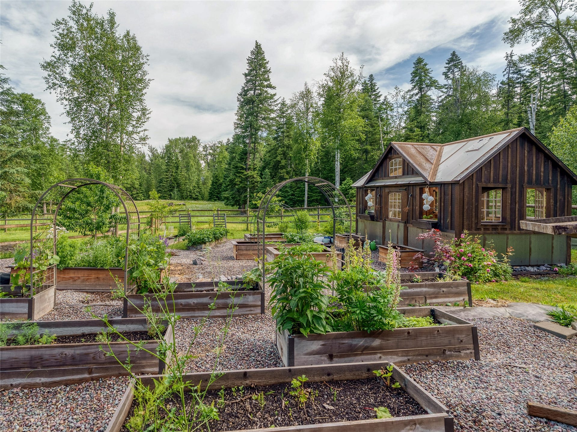 28012 Meadow Creek Road, Bigfork, MT 59911 Charming garden compound featuring raised bed vegetable gardens with mature landscape plantings, gravel pathways, and a dark wood-finish cottage structure with metal roofing