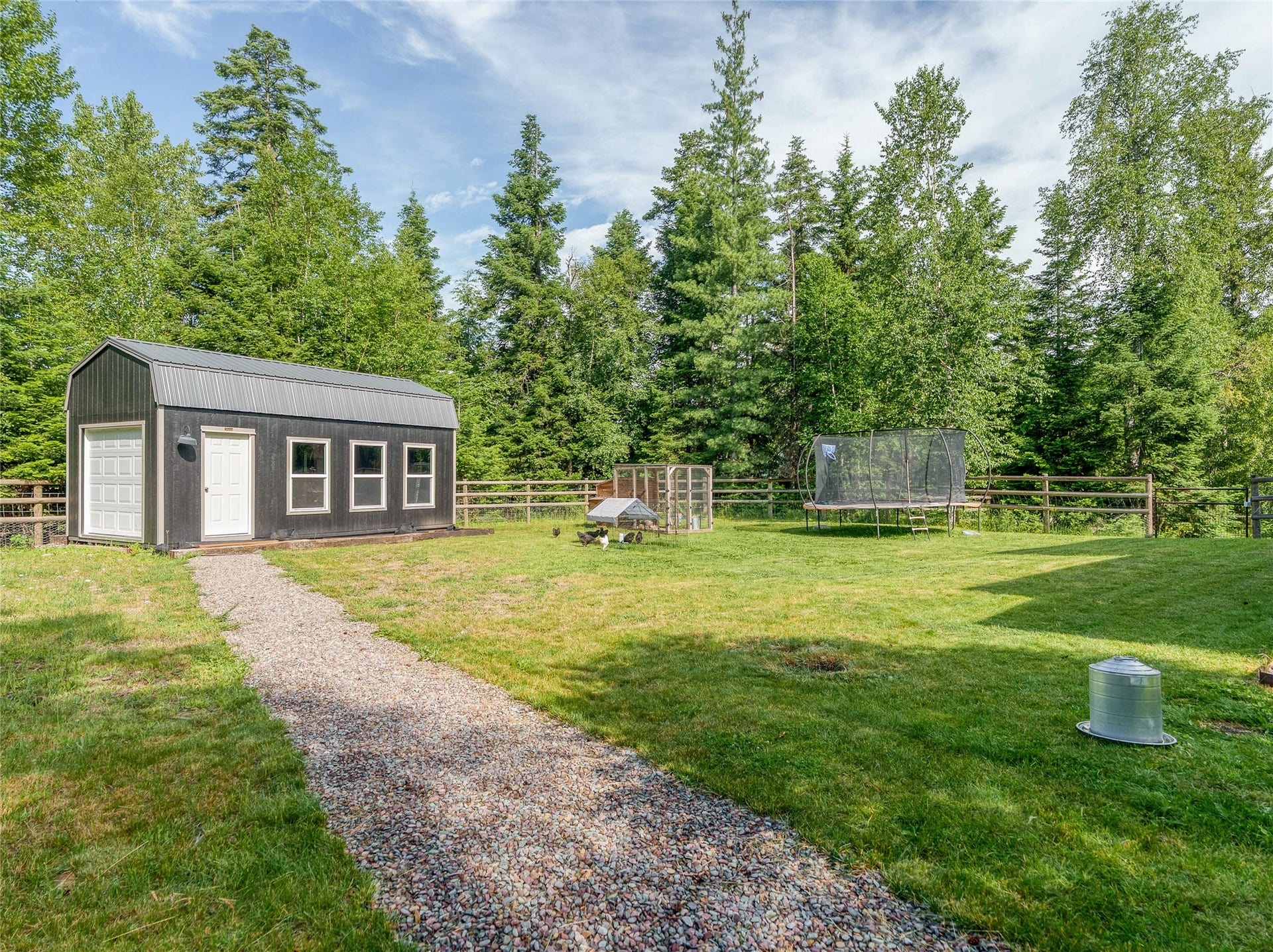 28012 Meadow Creek Road, Bigfork, MT 59911 Spacious rural property featuring a charcoal-finish storage barn with dual entry doors and multiple windows, gravel driveway approach, fenced paddocks, and mature evergreen and deciduous tree backdrop