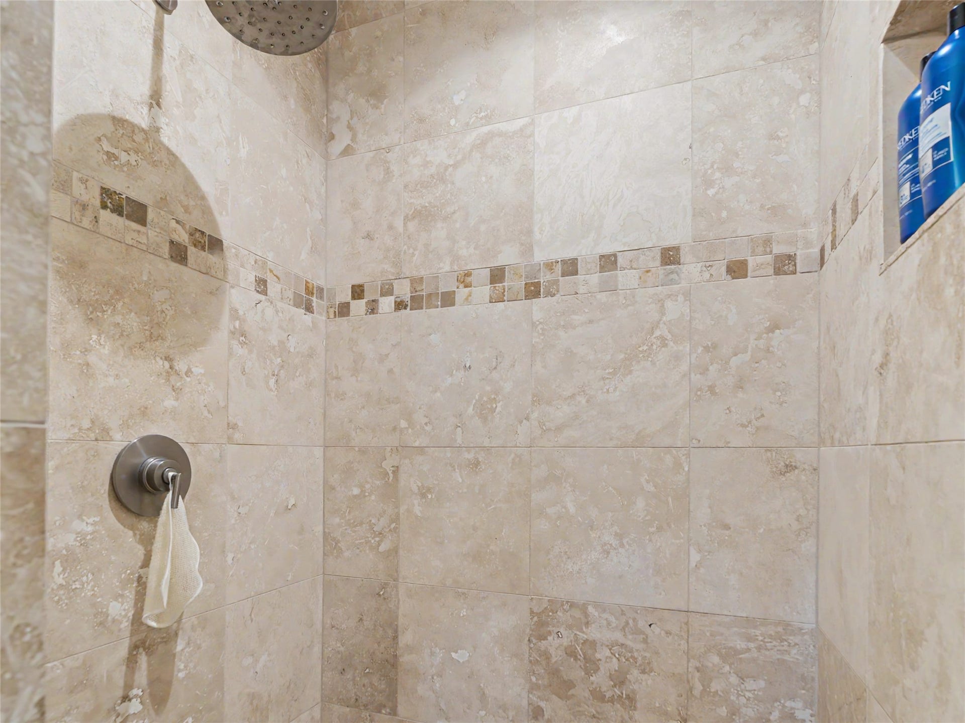 28012 Meadow Creek Road, Bigfork, MT 59911 2nd bathroom shower enclosure featuring large format stone-finish wall tiling, a decorative mosaic tile accent band, a built-in shower niche, a rainfall showerhead, and a brushed metal shower control