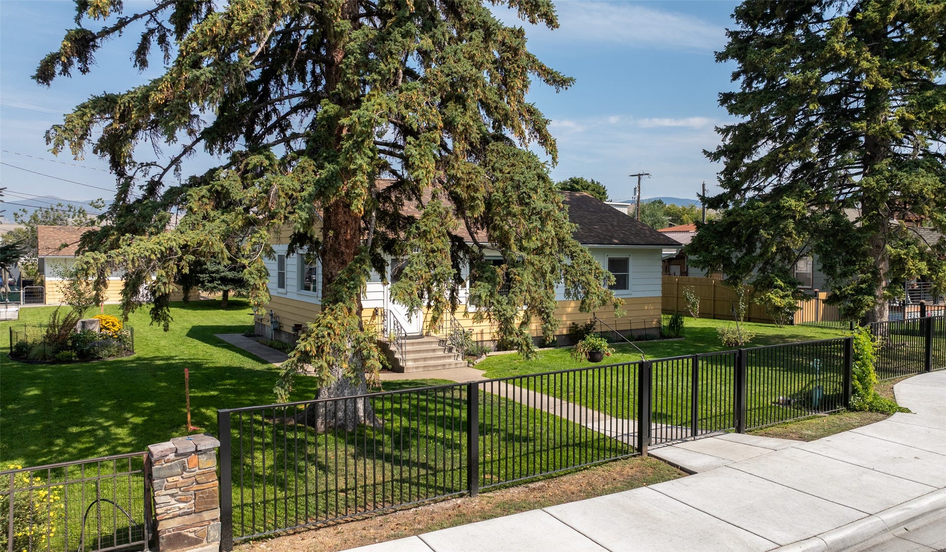 Manicured lawn with mature evergreen trees, stone-clad fence pillars, and a black wrought iron fence