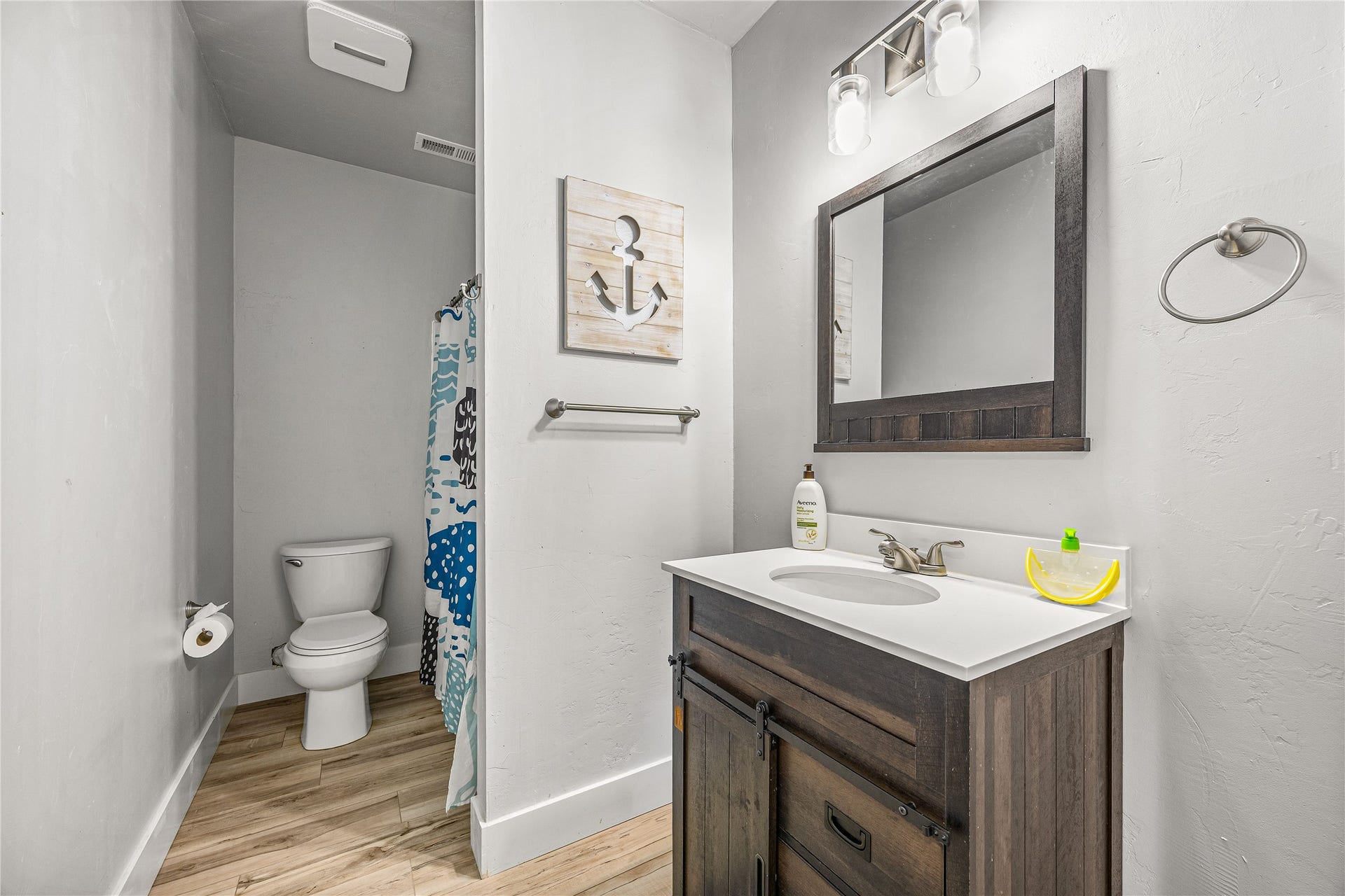 Wood-finish flooring with a white toilet, dark wood-finish vanity, single basin sink, and a framed mirror with overhead lighting