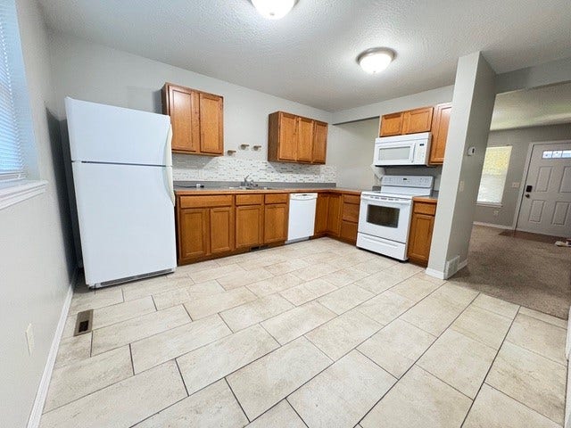 Kitchen featuring light wood-finish cabinetry, white appliances, light-toned tiled flooring, and a patterned tile backsplash
