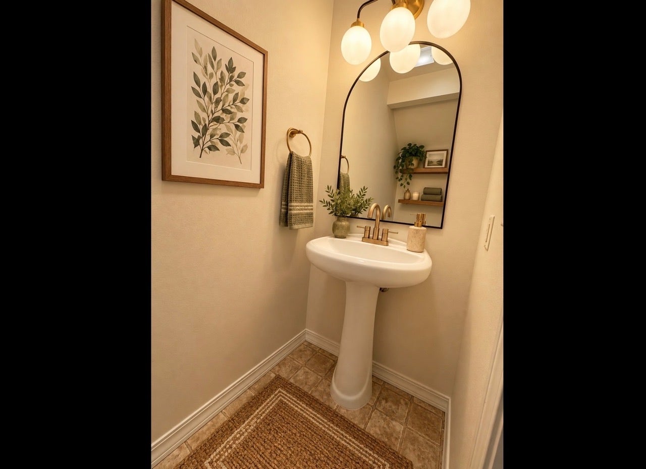 Pedestal sink with gold-tone fixtures, arched mirror, wall-mounted light fixture, and tile flooring