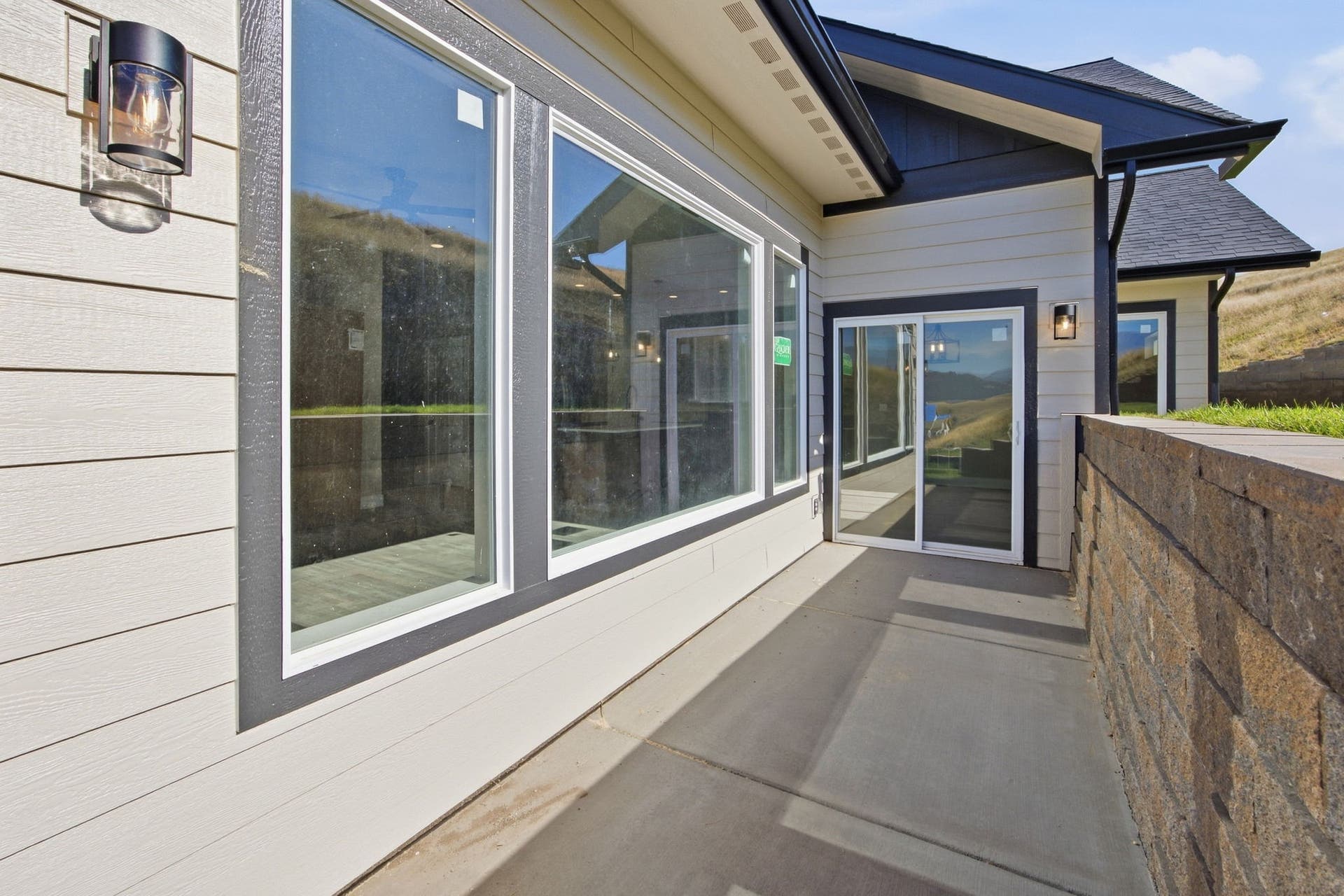 Exterior siding with contrasting trim, multiple large windows, sliding glass door, concrete patio, and a stone retaining wall