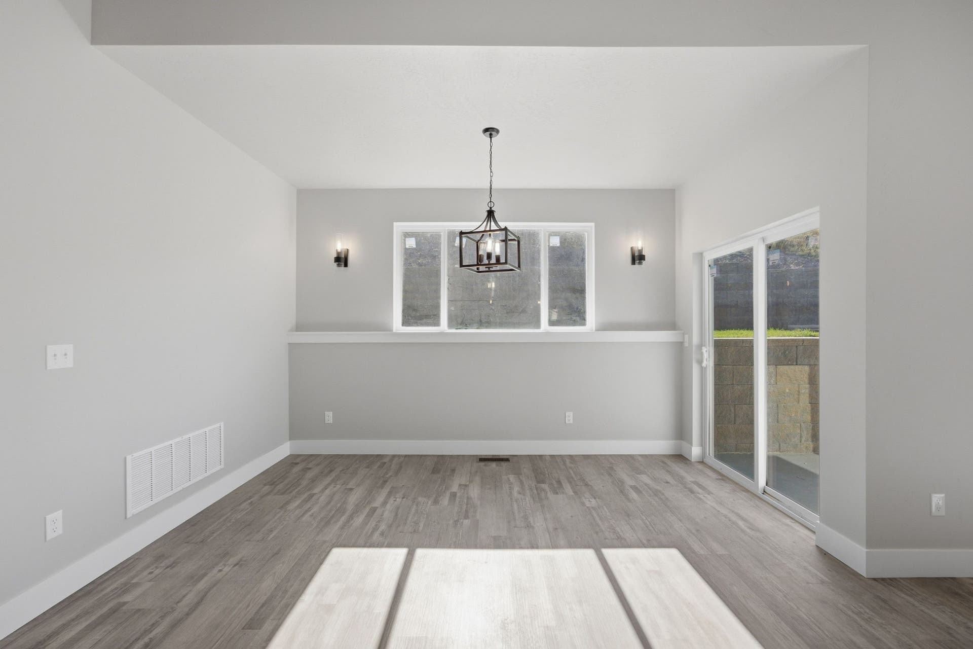 Spacious room featuring wood-finish flooring, a contemporary chandelier, two wall sconces, a large window, and a glass sliding door