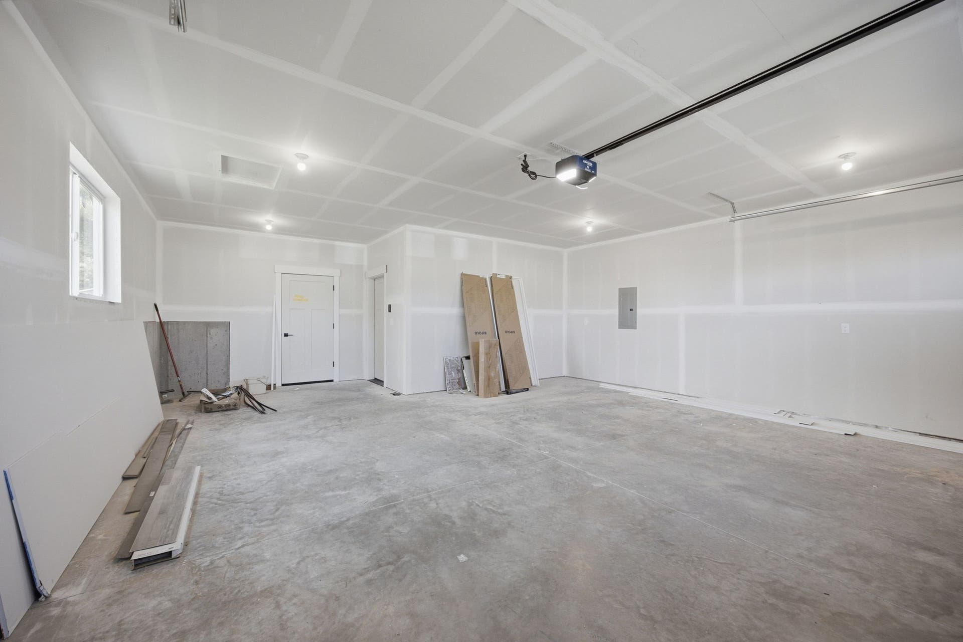 Spacious concrete-floor interior with drywall walls and ceiling, featuring recessed lighting, a white window, and an overhead garage door mechanism