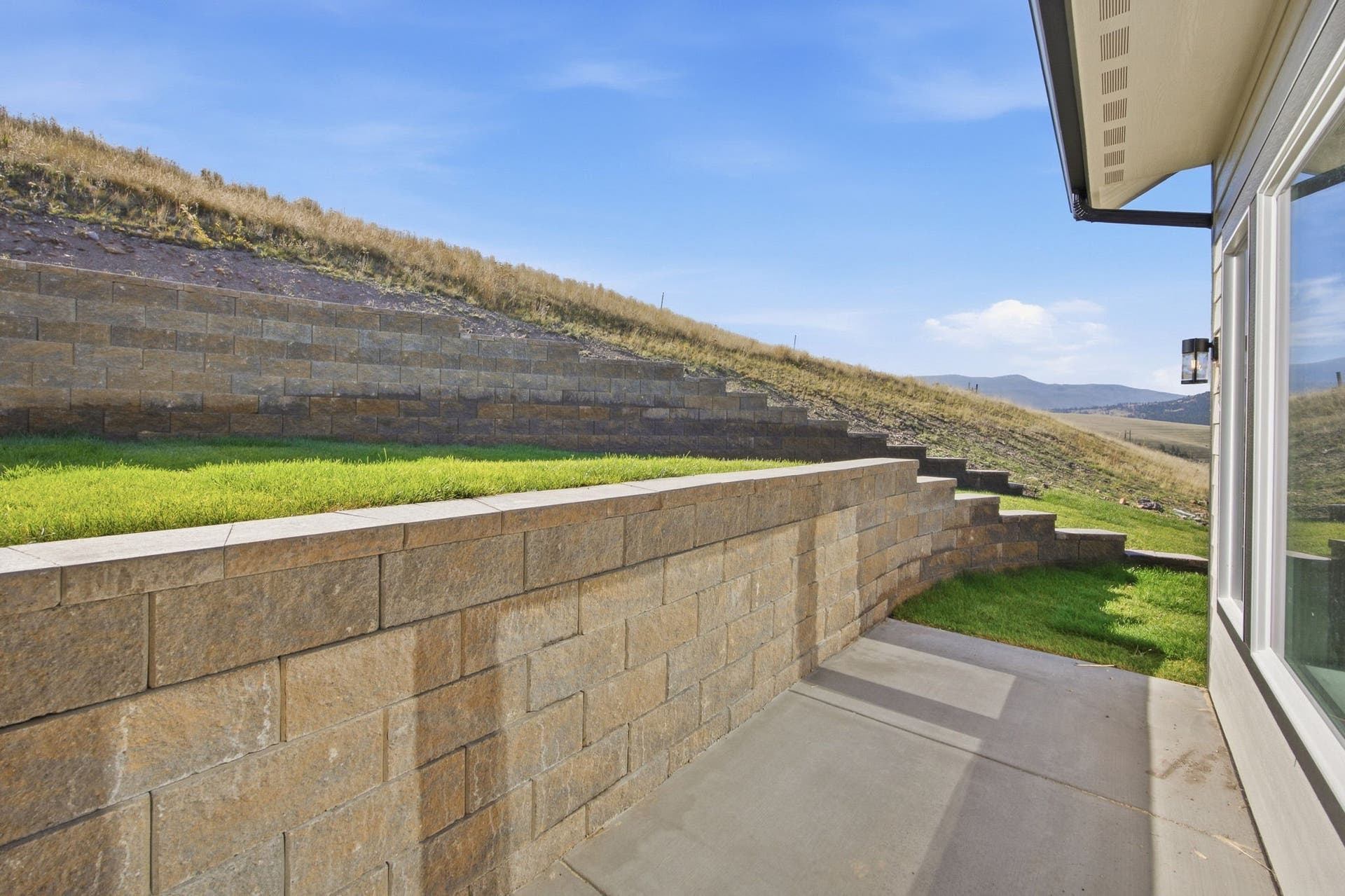 Concrete patio with a multi-tiered retaining wall, exterior wall sconce, and large window