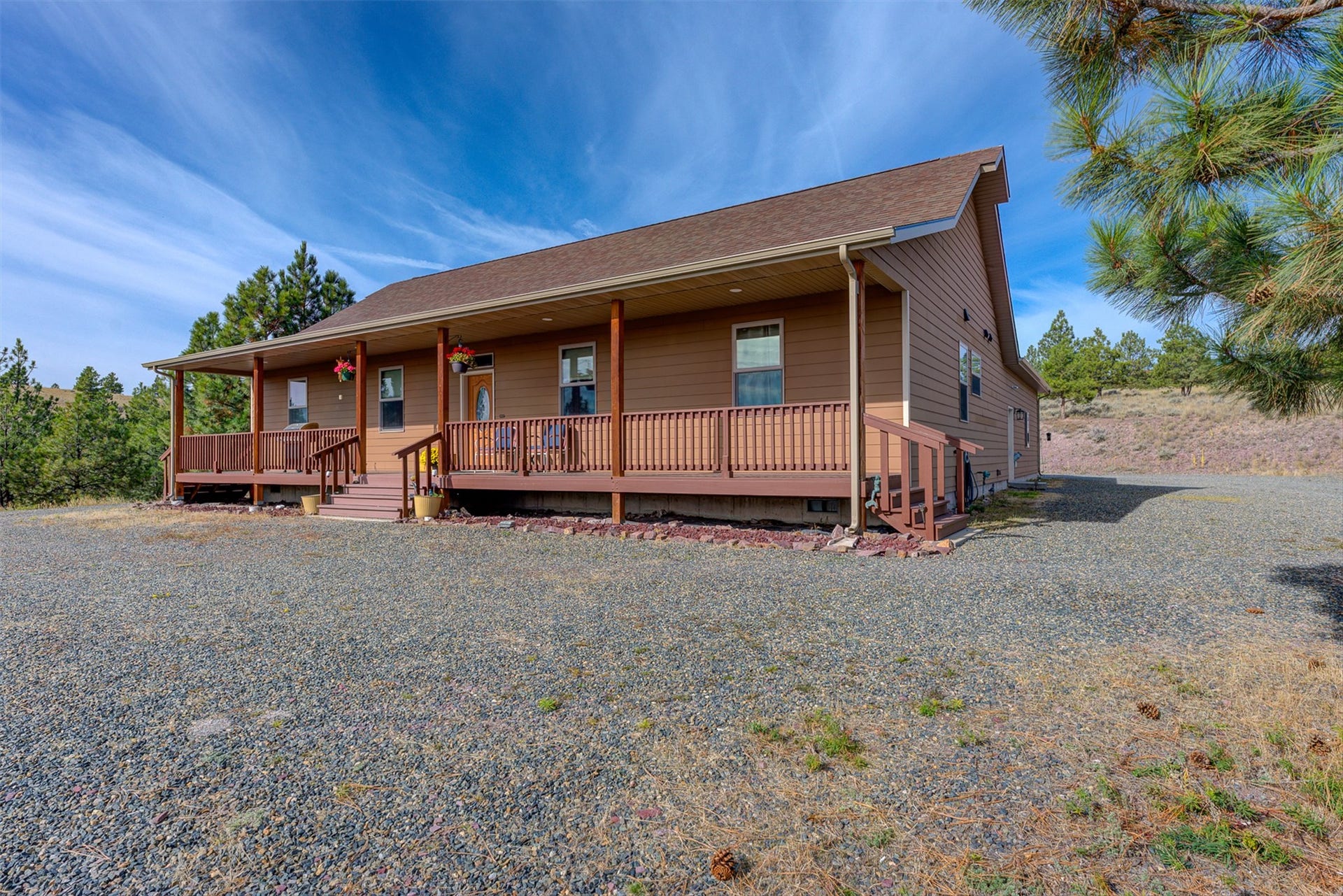 8224 Hillside Drive, Helena, MT 59602 Full-length covered porch, natural wood railing, light brown siding, brown asphalt shingle roof, and a gravel surface