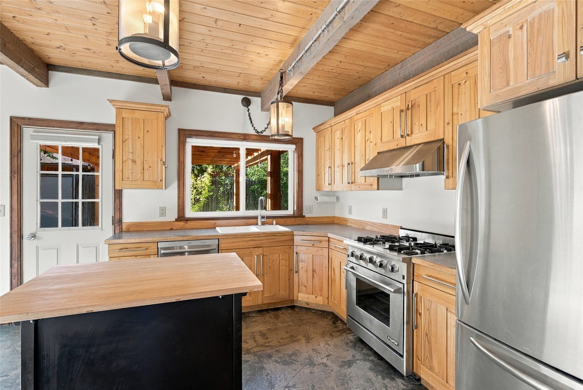Wood-finish cabinetry, stainless steel appliances, exposed wood beam ceiling, and a large window above the sink