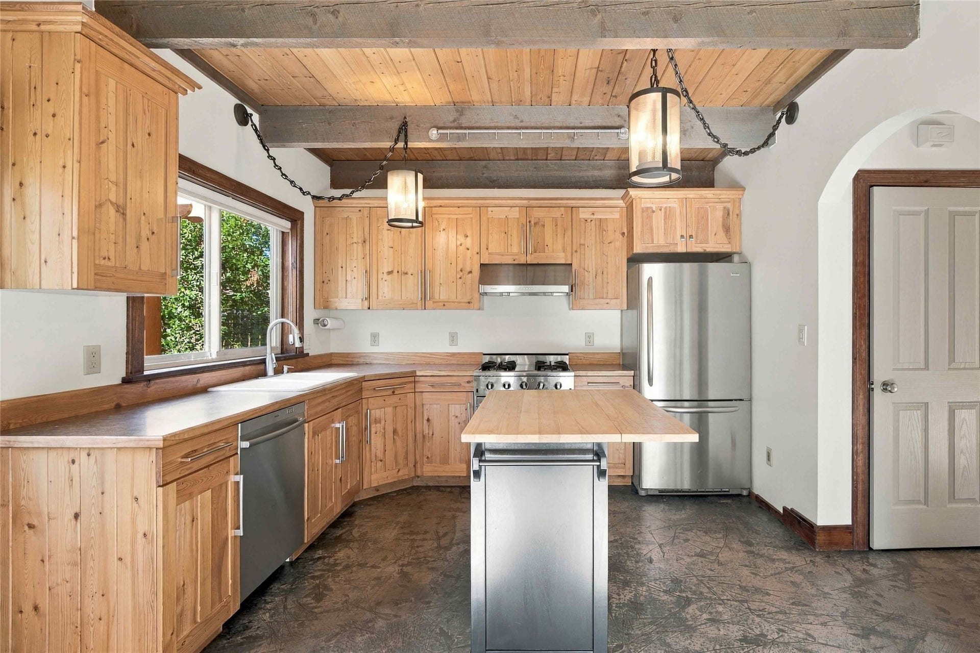 Exposed wood beam ceiling with wood paneling, wood-finish cabinetry, stainless steel appliances, central island with wood countertop, and concrete flooring