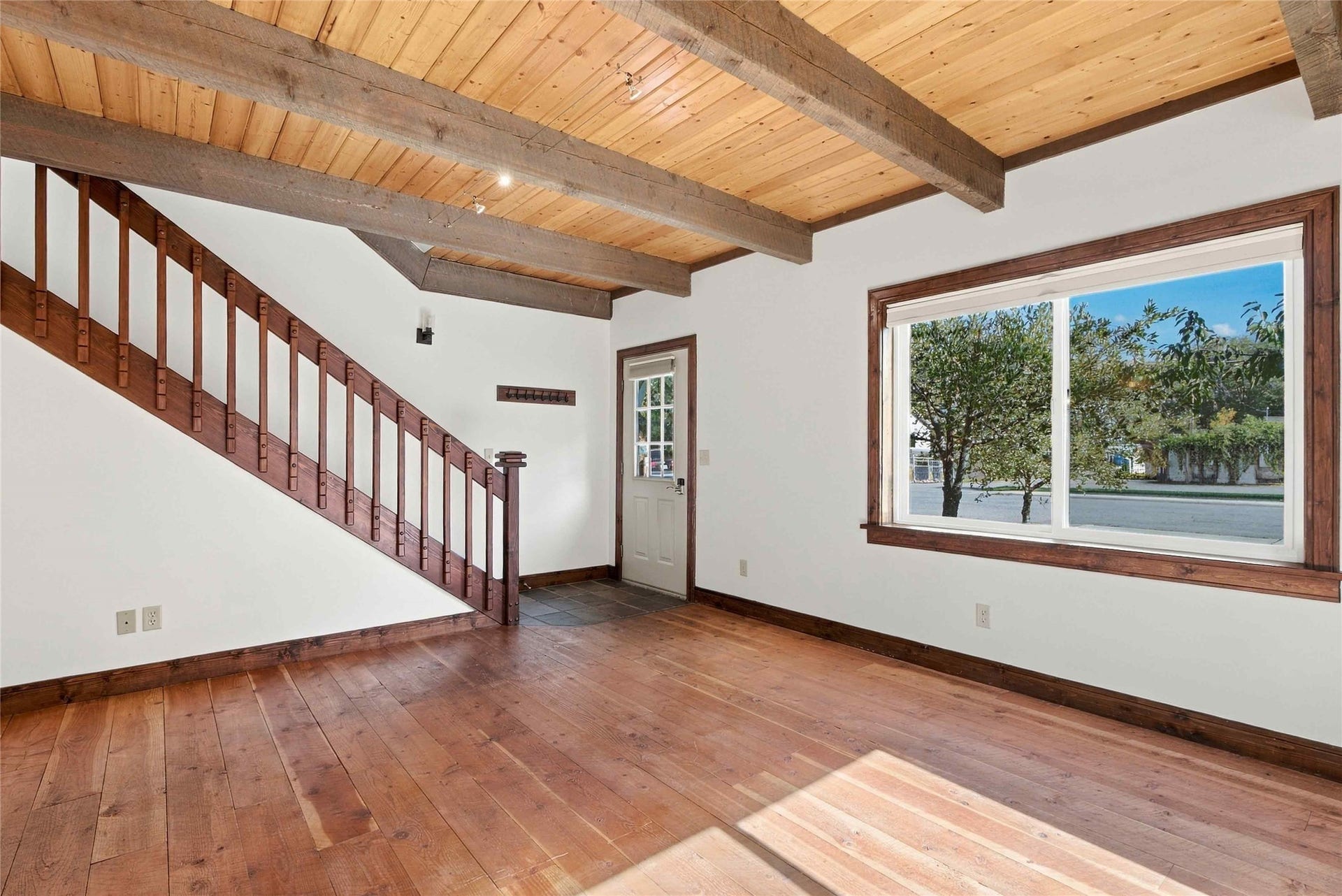 Exposed wood beam ceiling, wood-finish flooring, wood staircase with balusters, wood-trimmed window, and front entry door with glass panels