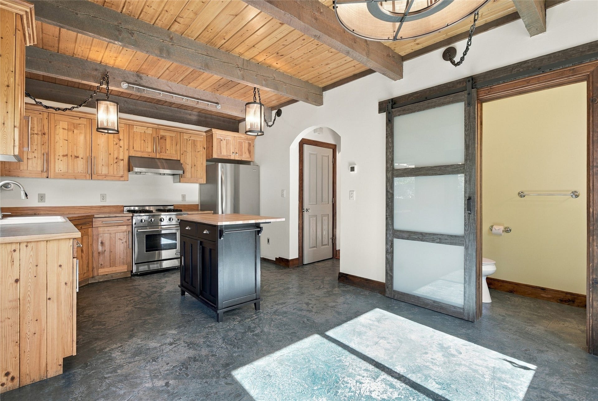 Exposed wooden beams and ceiling, stainless steel appliances, central kitchen island, wood-finish cabinetry, and a sliding barn door with frosted glass panels