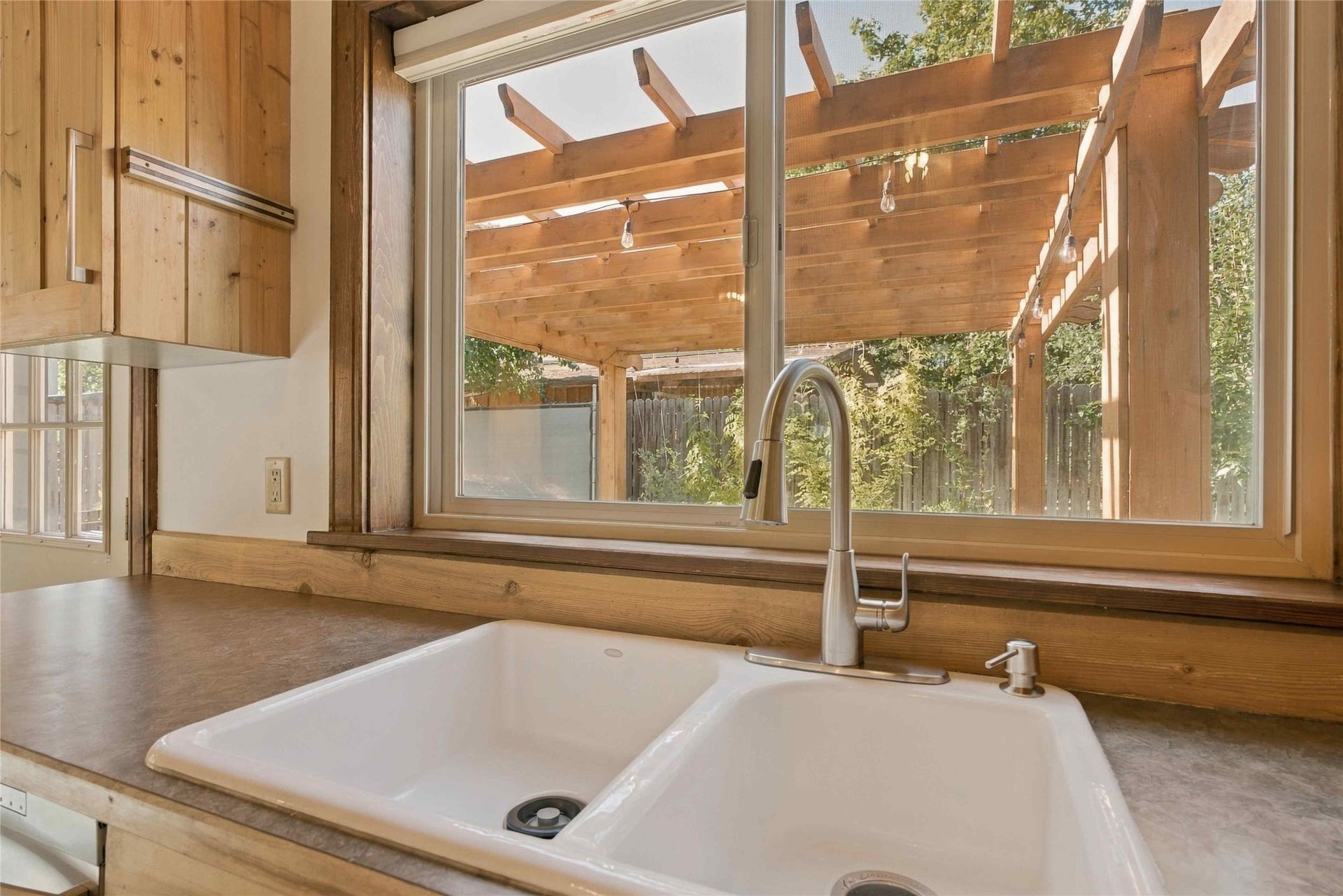 Double basin sink with a high-arch faucet, set into a dark countertop, framed by natural wood cabinetry and window trim