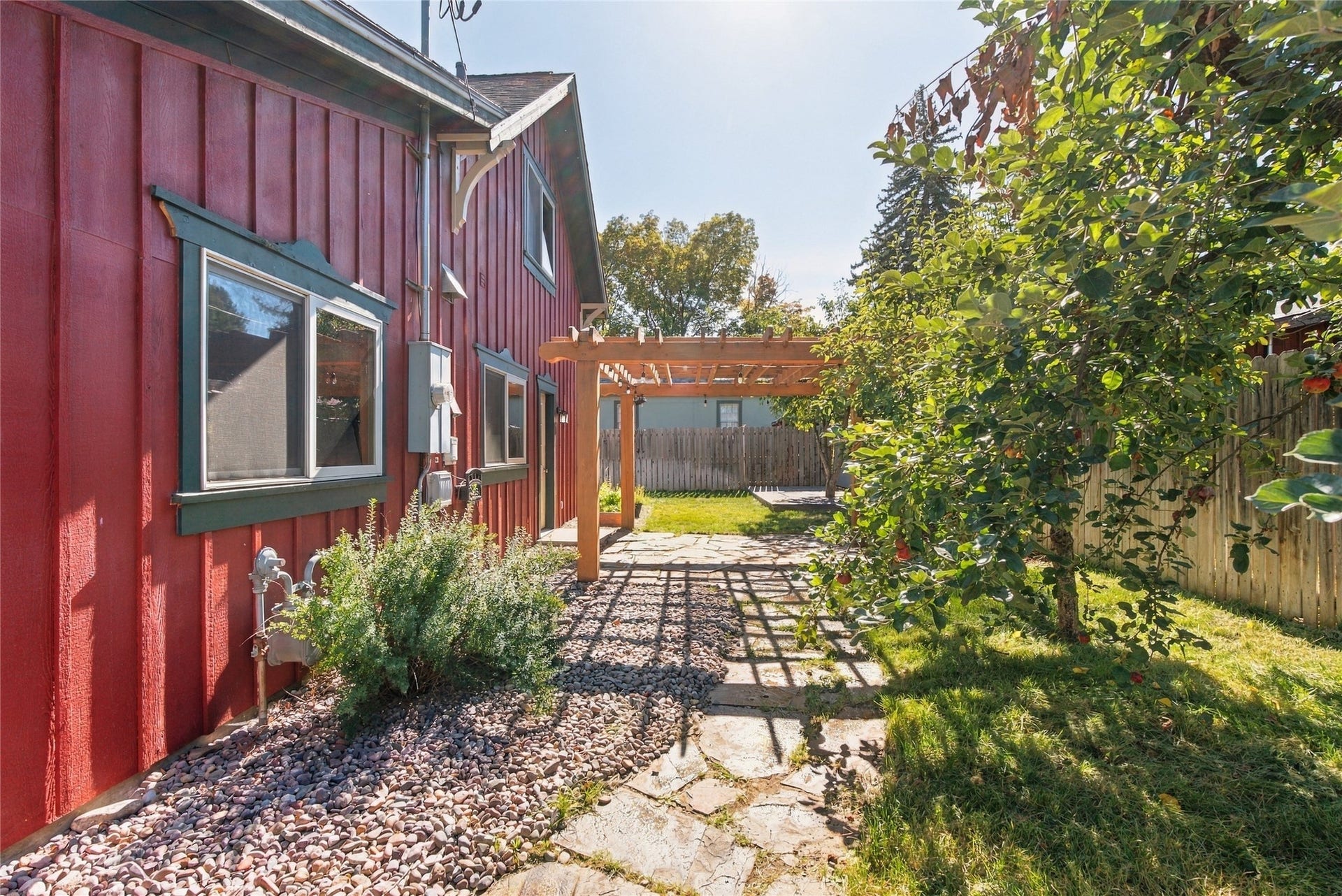 Red vertical siding with dark green trim, stone paver pathway, gravel landscaping, wood pergola, and established trees