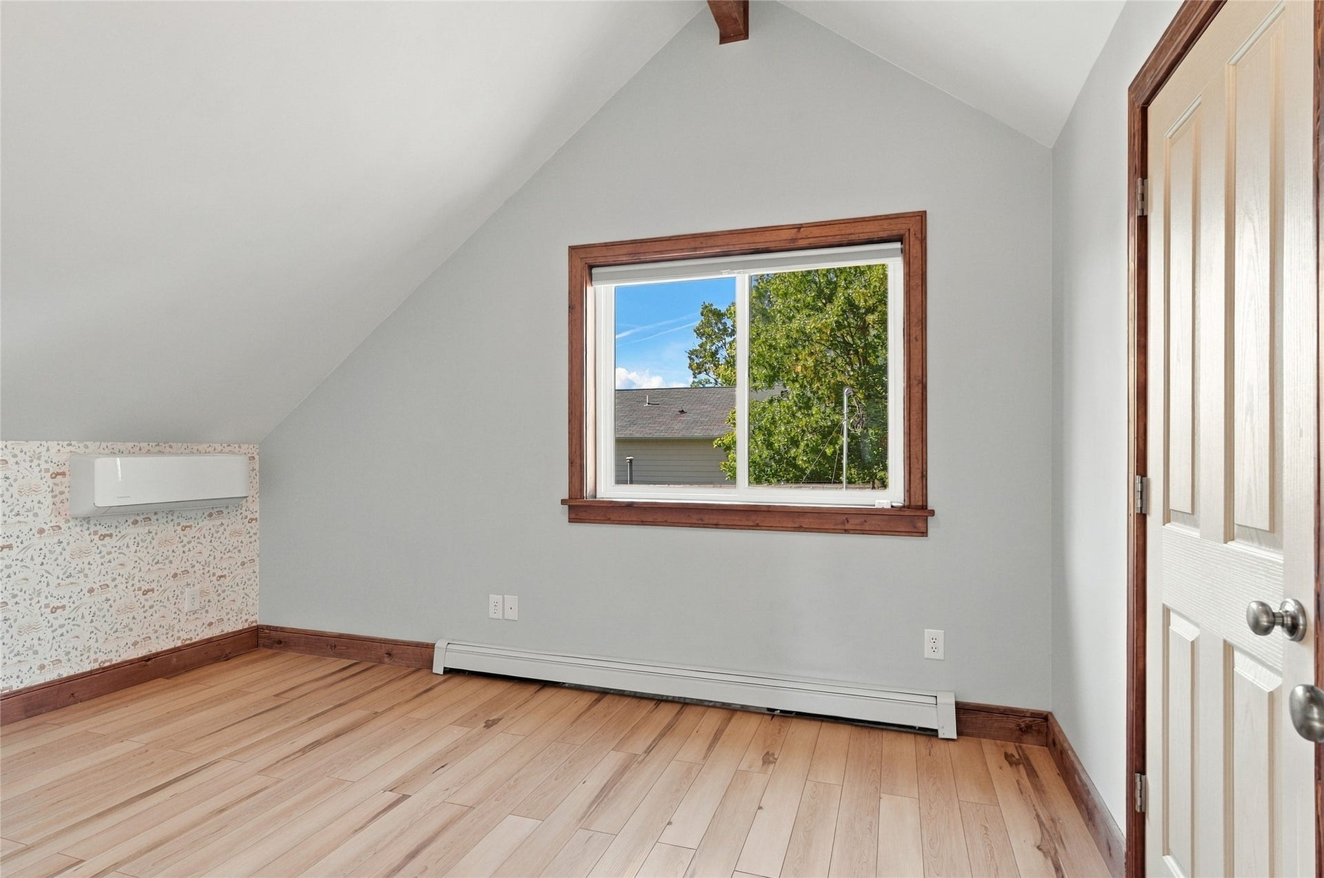 Wood-finish flooring, accent wall with patterned wallpaper, built-in wall unit, wood trim around window and door, and an angled ceiling