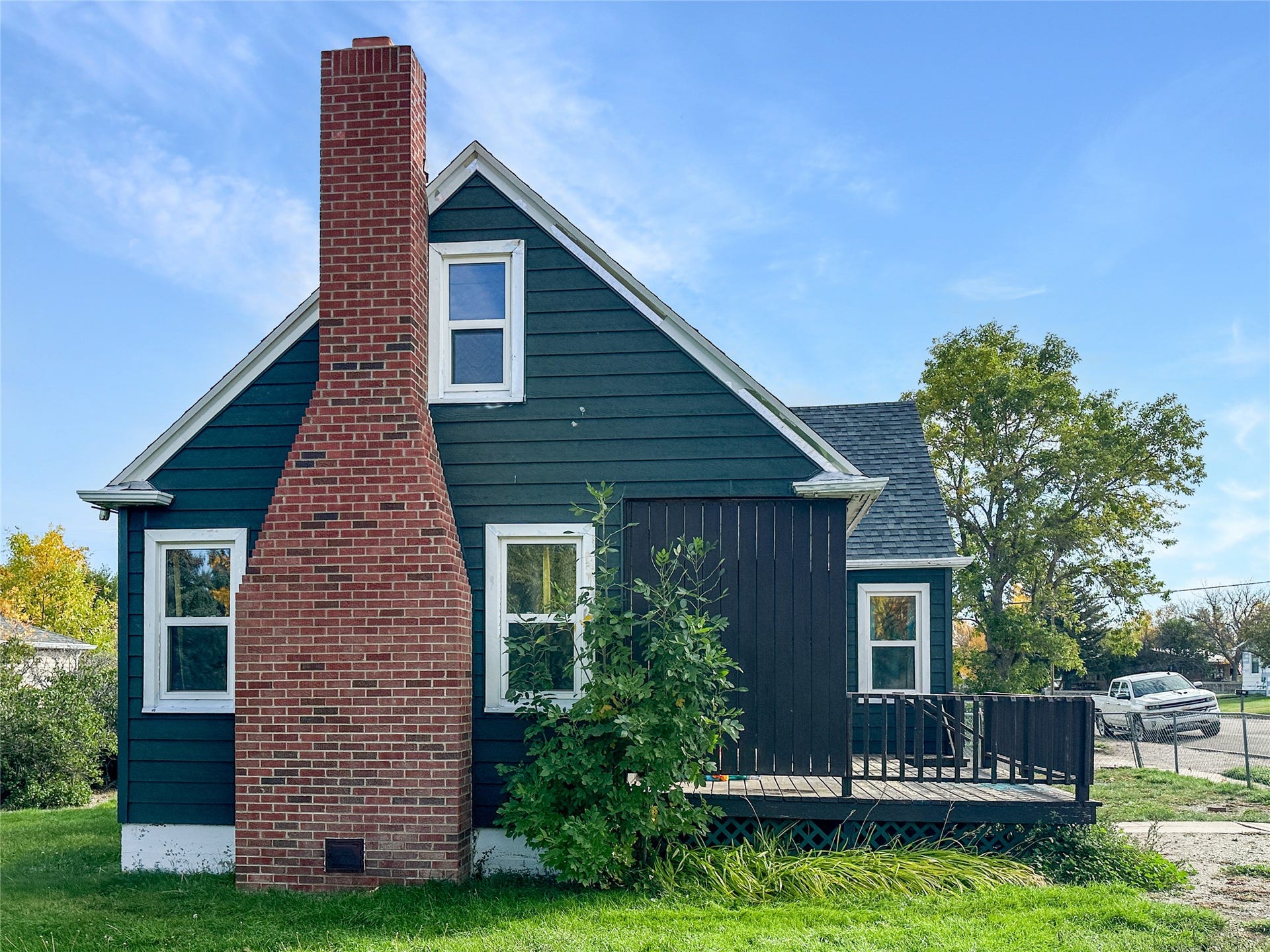 1123 2nd Street N, Shelby, MT 59474 Exterior featuring dark blue siding, a prominent brick chimney, white window trim, and a dark-stained wood deck