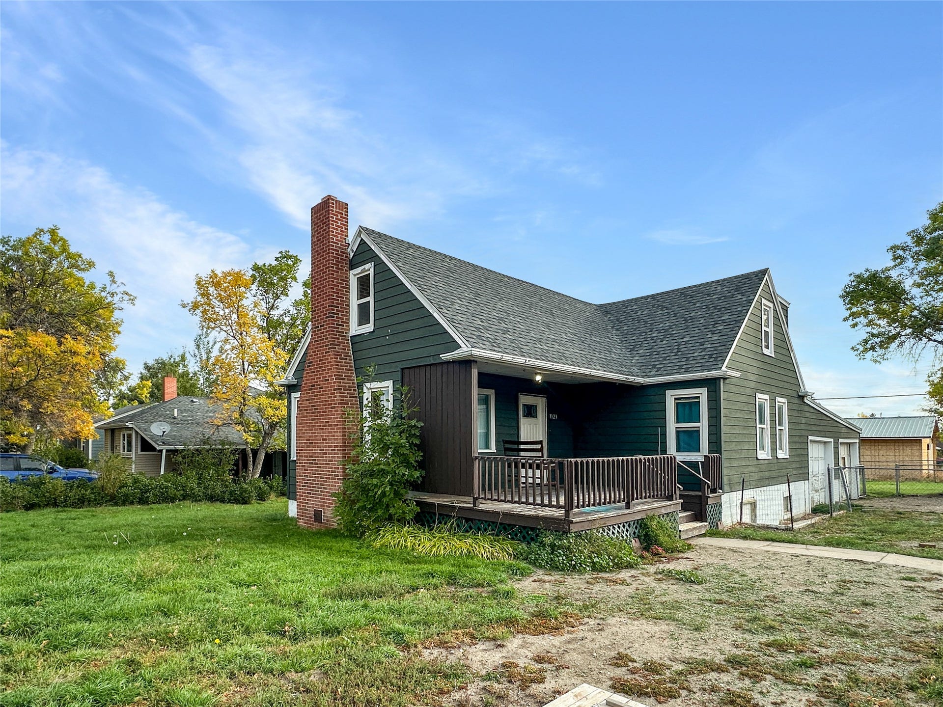 1123 2nd Street N, Shelby, MT 59474 Gabled roof residence featuring dark green siding, a prominent brick chimney, and a covered front porch with a wood railing