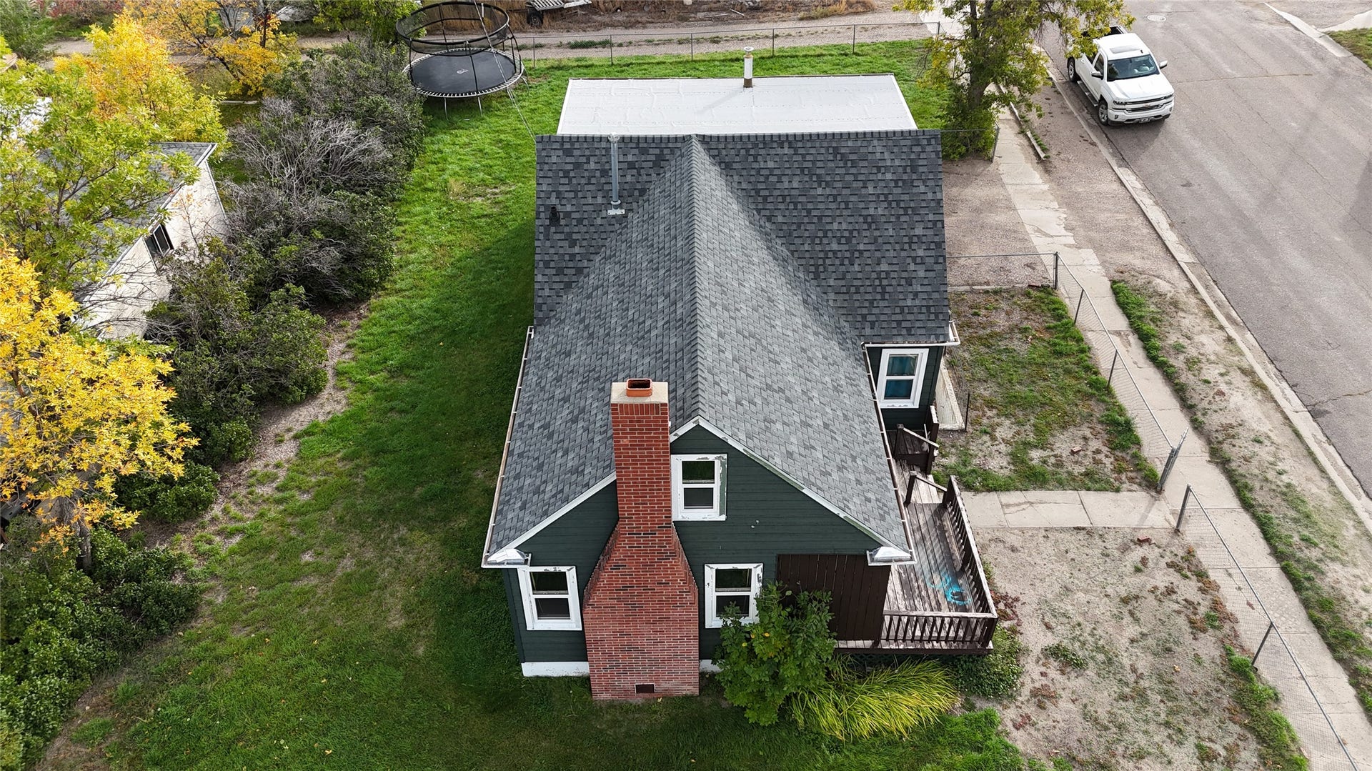 1123 2nd Street N, Shelby, MT 59474 Dark shingle roof, dark green siding, red brick chimney, white trim windows, and a wooden deck