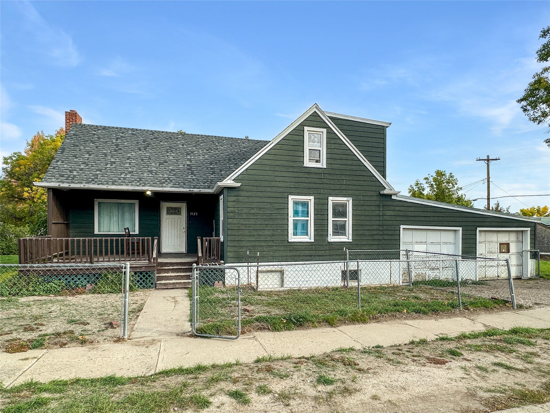 1123 2nd Street N, Shelby, MT 59474 Green siding, multi-pane windows, front porch with wood railing, attached garage, and a chain-link fence