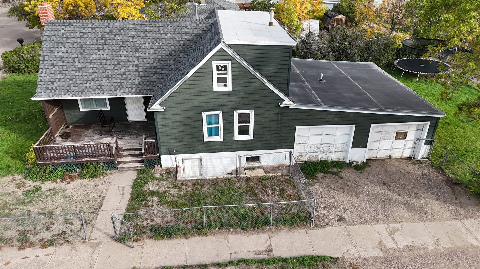 1123 2nd Street N, Shelby, MT 59474 Gabled roofline on half of house. Flat rolled roof upstairs, black rolled roof over garage, dark green siding, covered front porch with steps, attached two-car garage, and chain link fencing