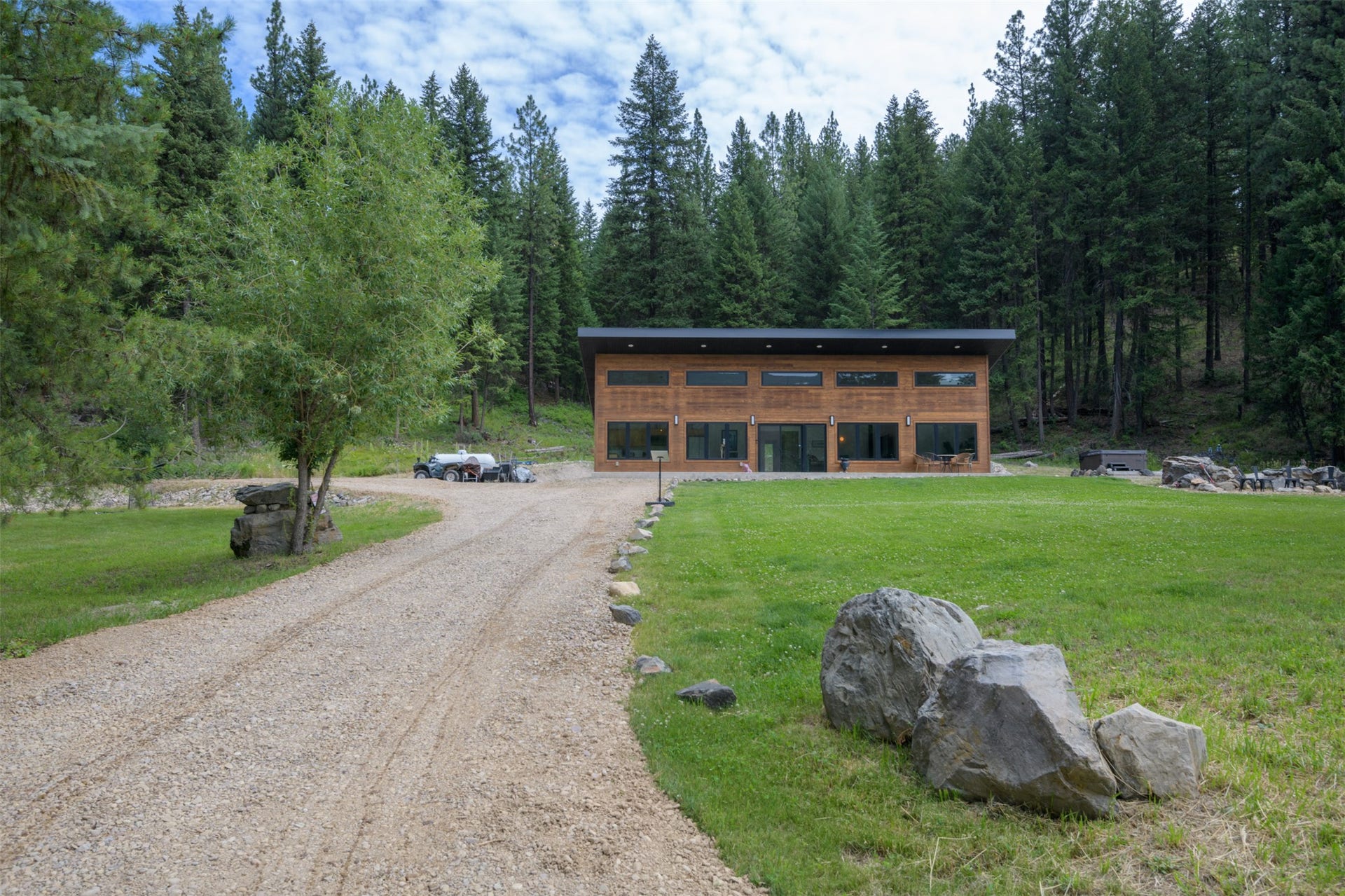 Wood-clad exterior with large windows, gravel driveway, expansive lawn, and mature trees