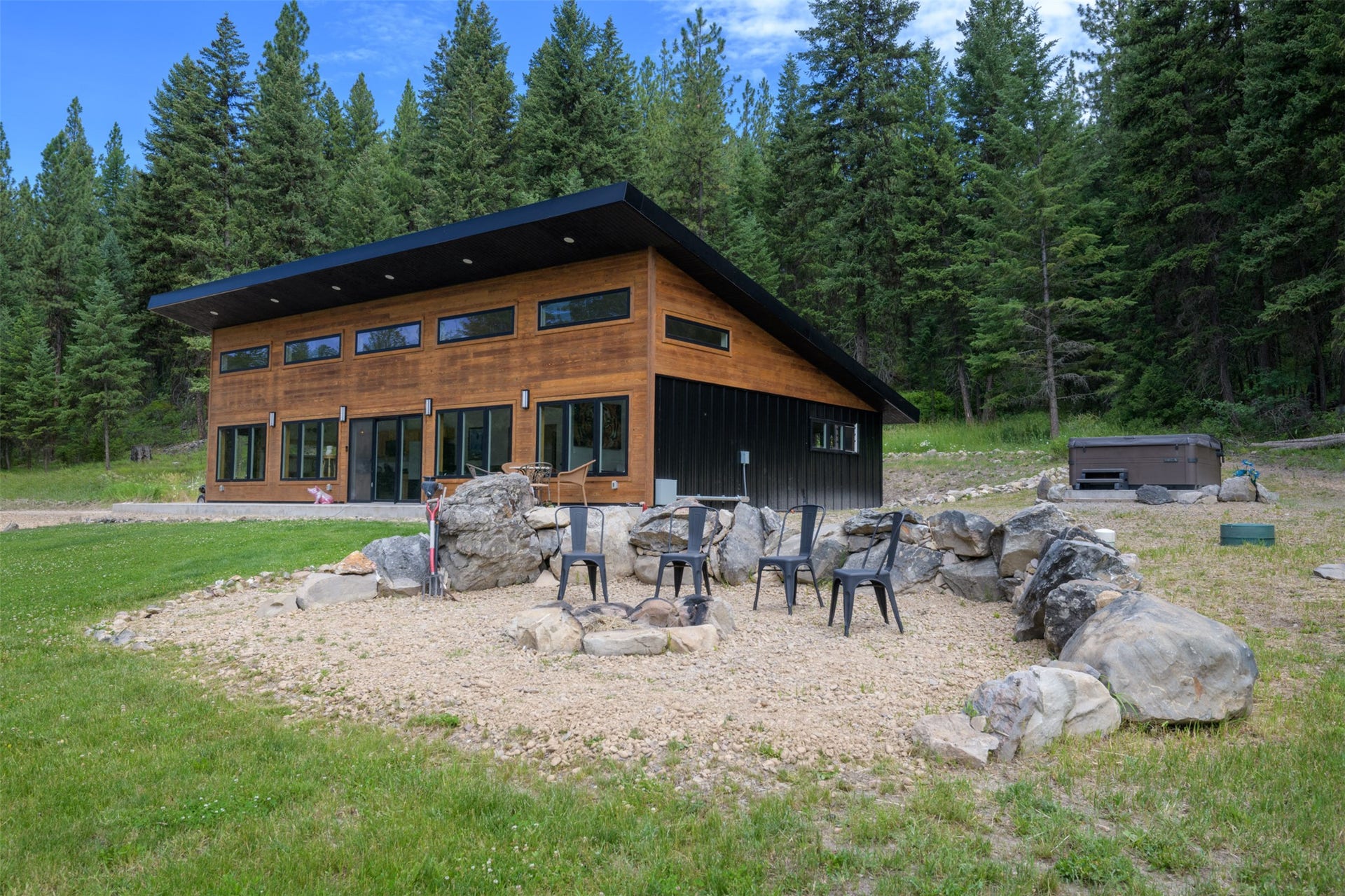 Modern exterior with wood siding and black trim, featuring a large outdoor fire pit area with stone seating, multiple sliding glass doors, and a built-in hot tub