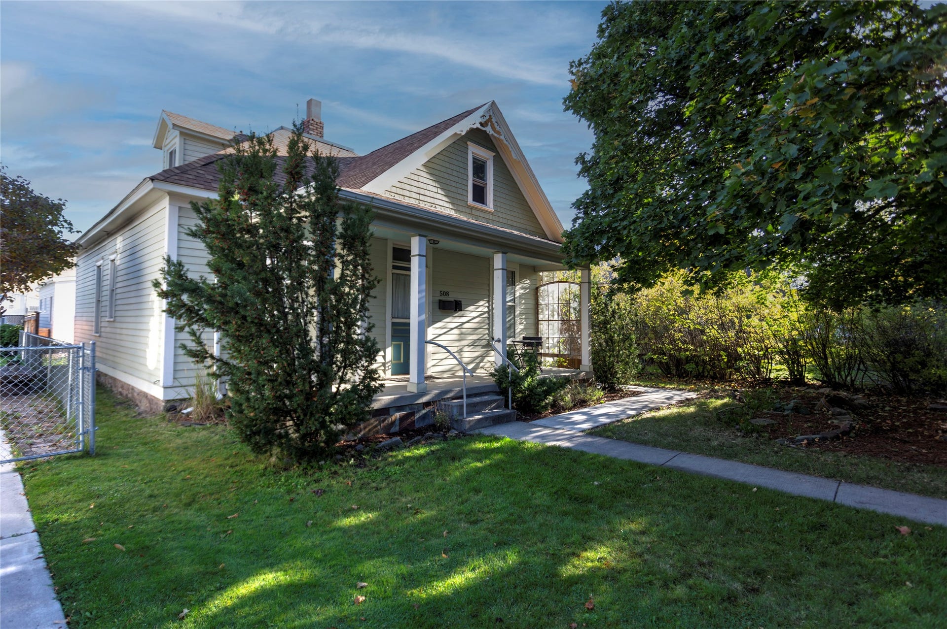 Gabled dormer, covered front porch with columns, front walkway, established trees, and chain-link fencing