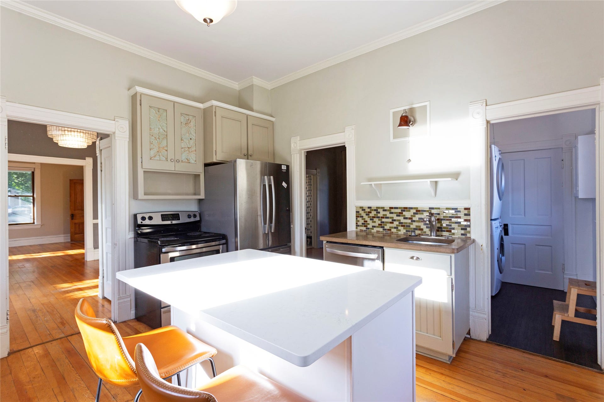 Wood-finish flooring, white island with countertop, stainless steel appliances, mosaic tile backsplash, and decorative cabinetry