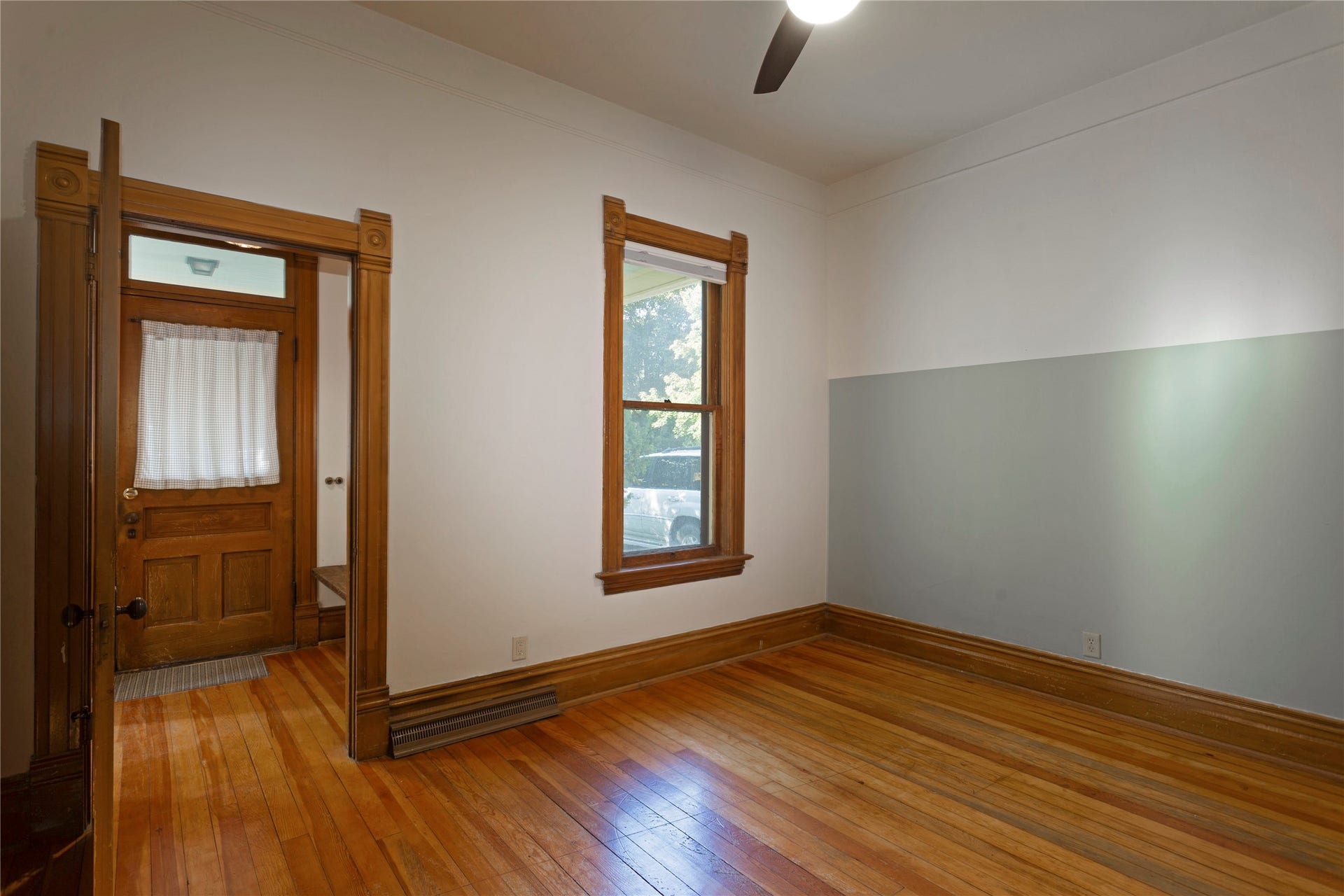 Hardwood flooring throughout, detailed wood trim, window with wood frame, ceiling fan fixture, and wood-paneled door with transom window