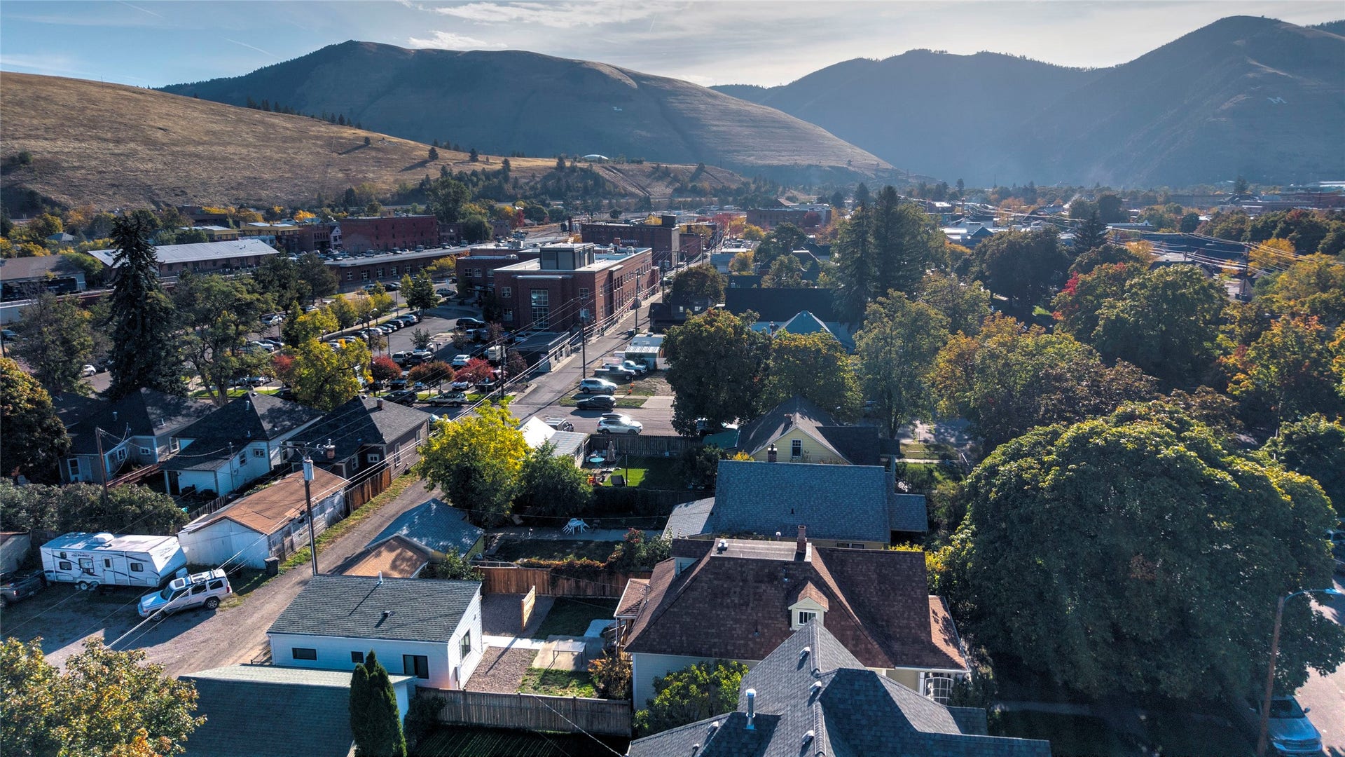Urban landscape featuring commercial and residential structures, abundant mature trees, and a distant mountain range