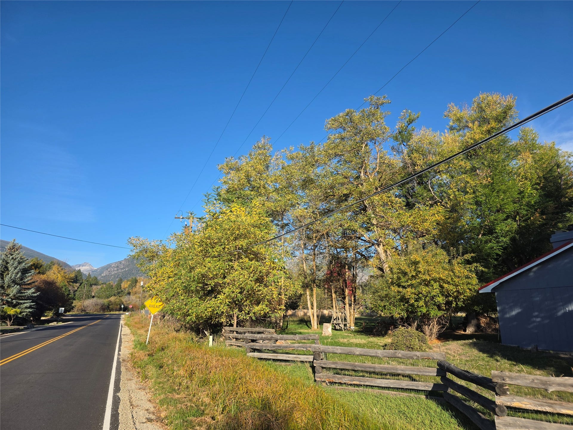 115 Ricketts Road, Hamilton, MT 59840 Mountain backdrop, mature trees, split-rail fencing, paved road frontage, partially visible structure with a gabled roof