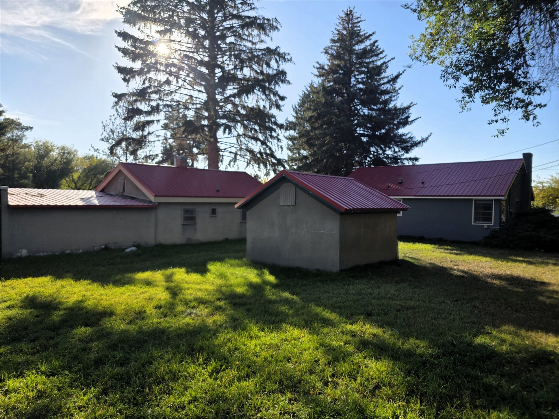 115 Ricketts Road, Hamilton, MT 59840 Expansive green lawn with mature trees, featuring a stucco-finish outbuilding with a red metal roof and a main structure with a red metal roof and grey siding
