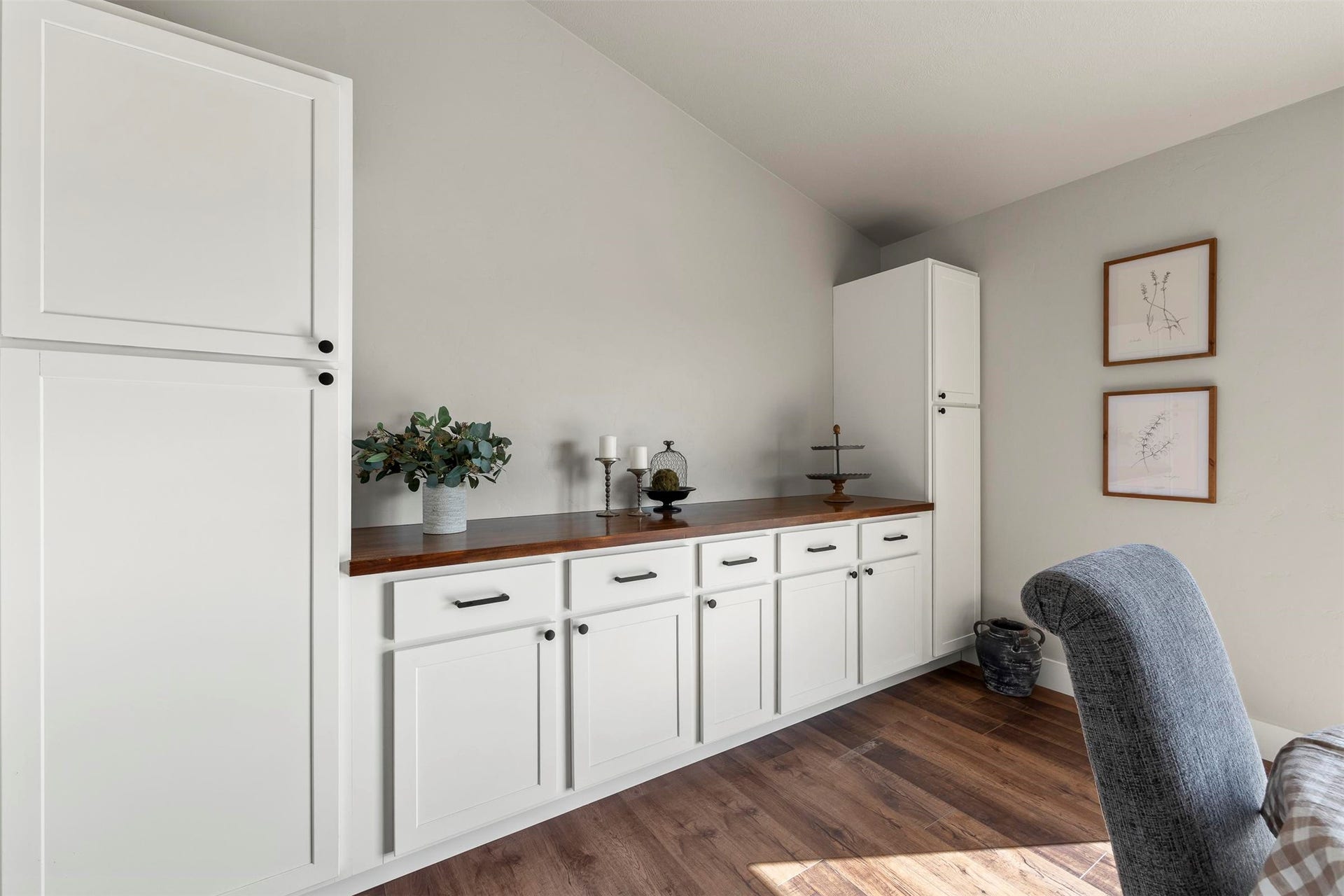 Extensive built-in cabinetry with white shaker-style doors, dark wood-finish countertop, black cabinet pulls, tall integrated pantry, and wood-finish flooring