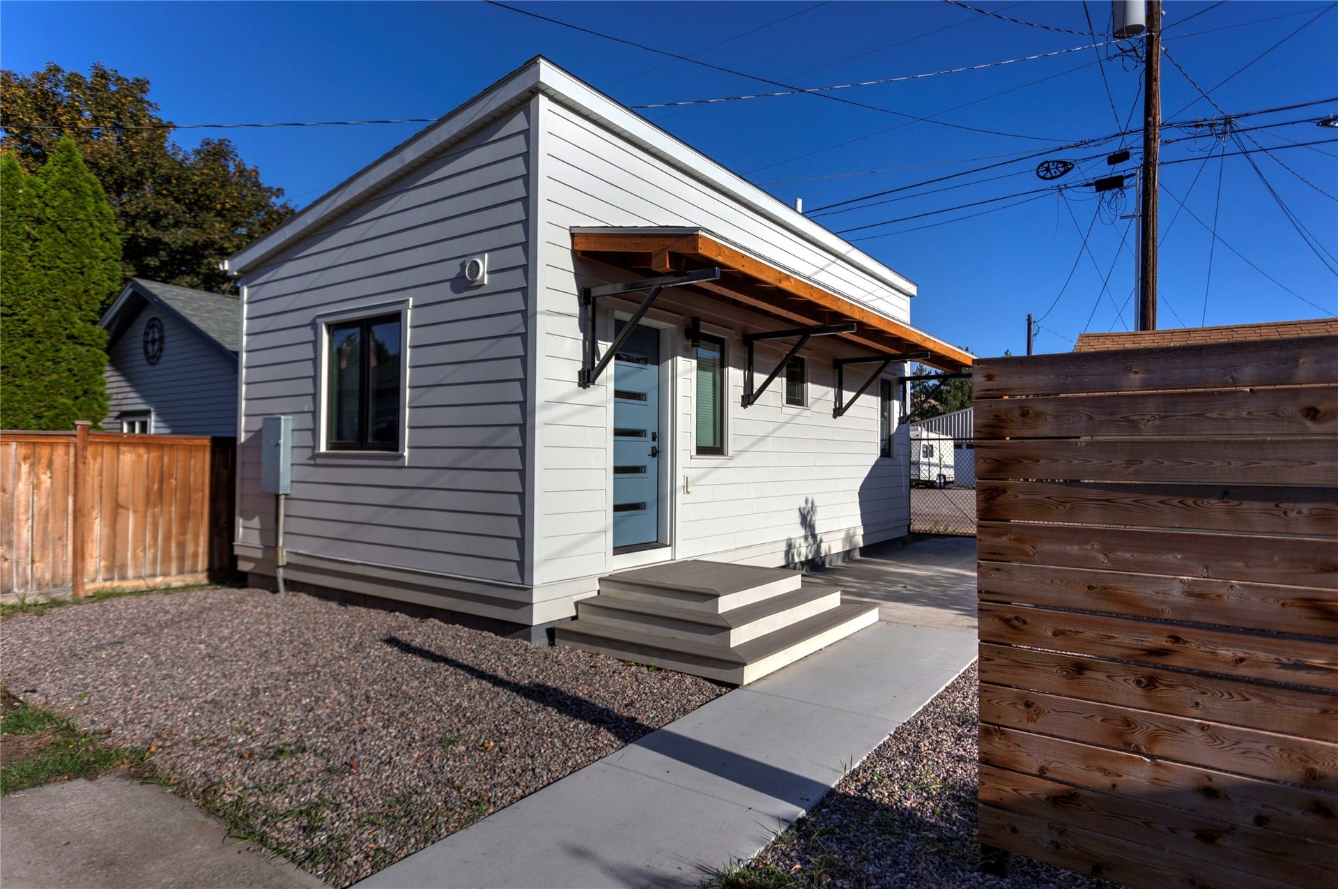 Modern exterior featuring horizontal siding, a light blue entry door, a wood-finish awning with black support brackets, a concrete pathway, and a wood privacy fence