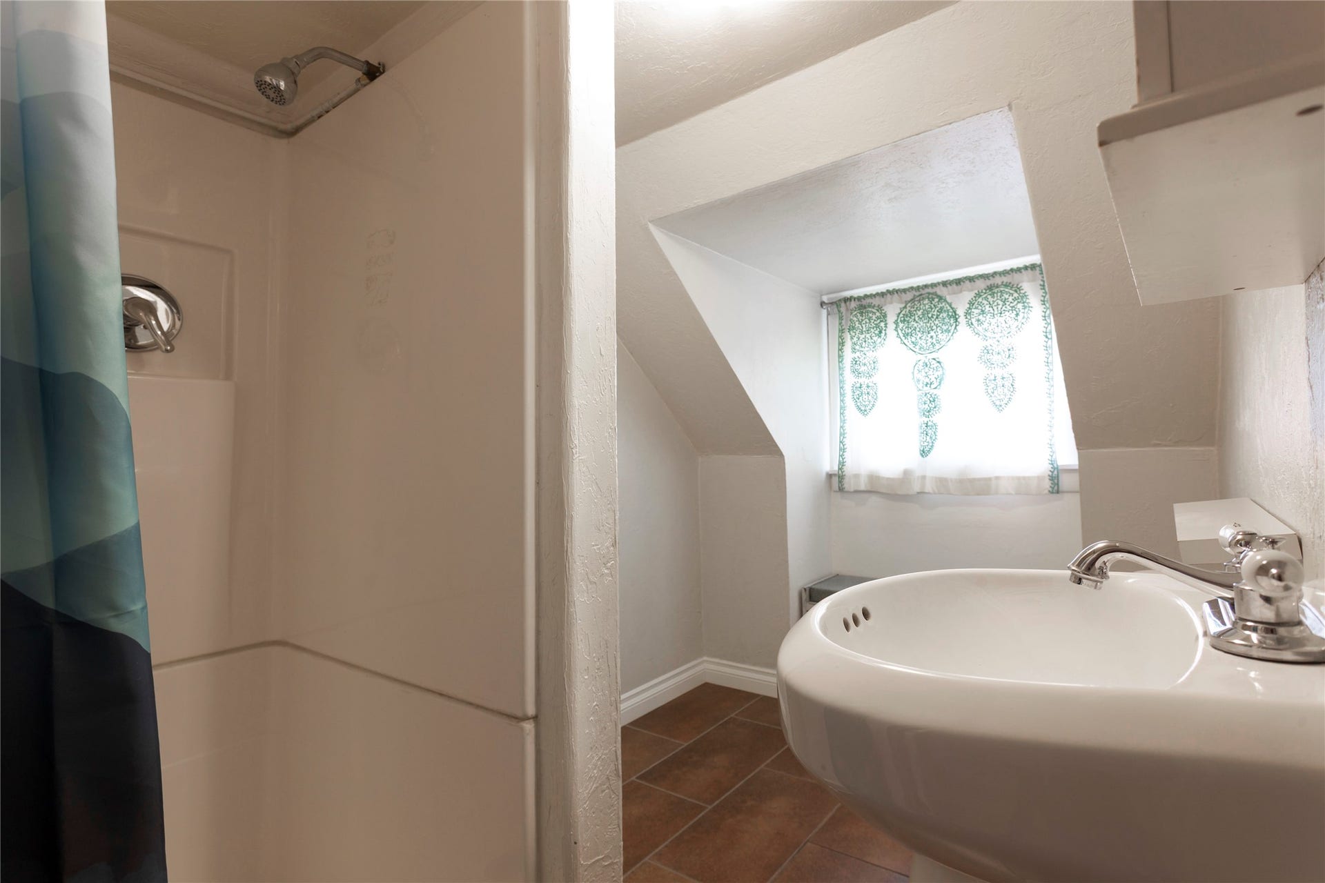 Pedestal sink with chrome faucet, white shower stall with chrome showerhead, and window with natural light