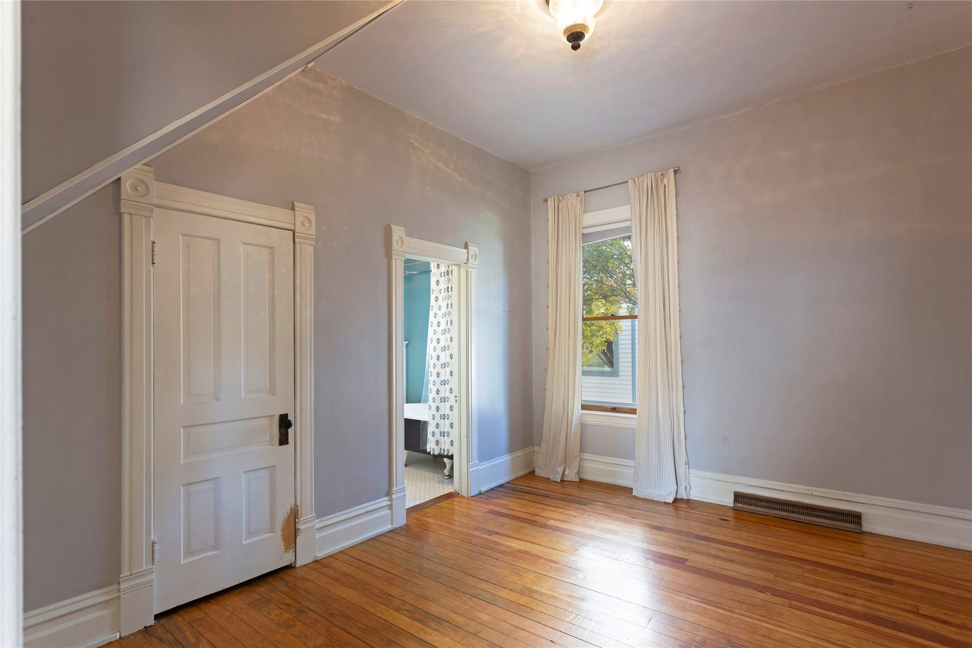Hardwood flooring, classic trim details, window with natural light, traditional ceiling light fixture, and an attached bathroom