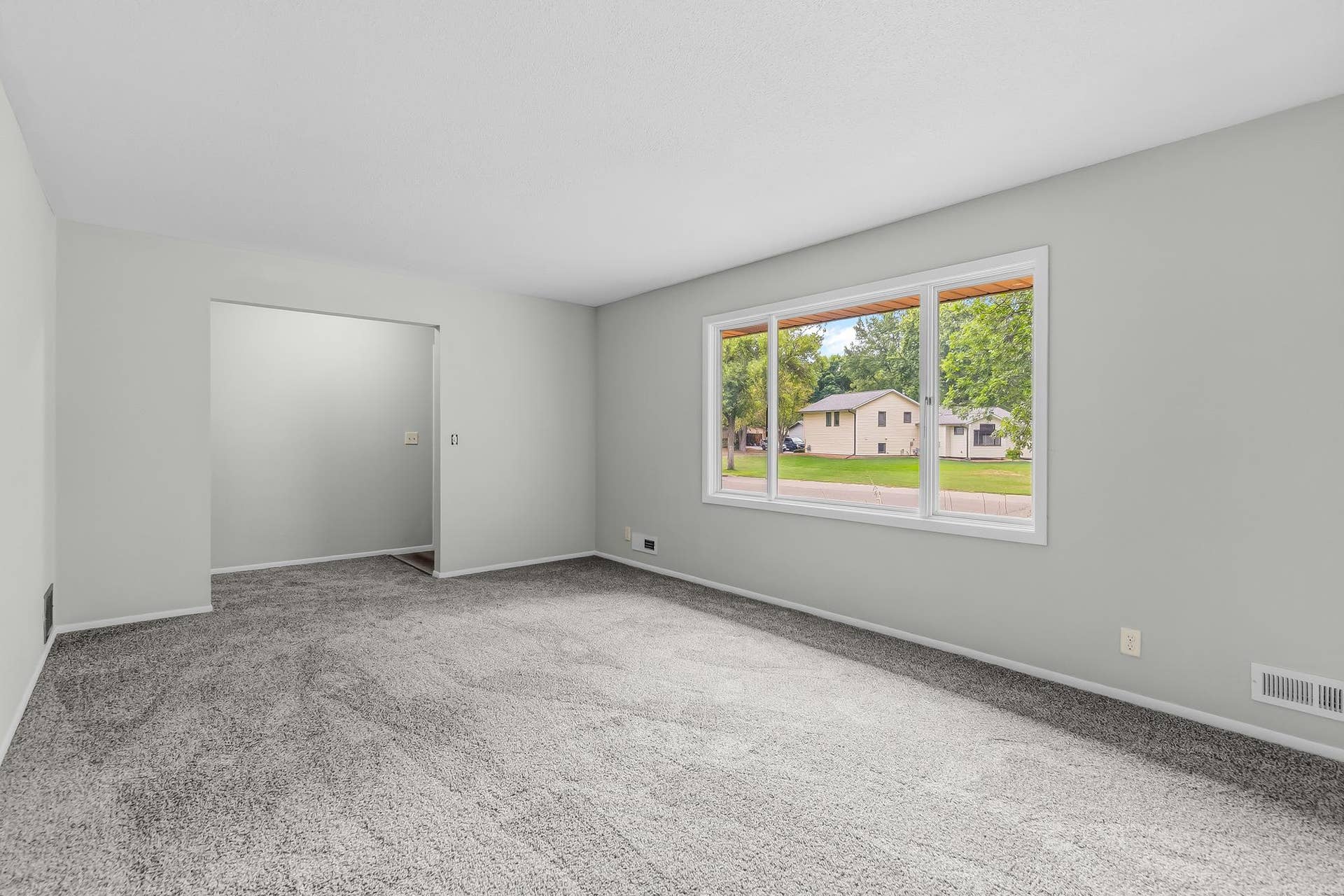 Spacious, light-filled living area with neutral finishes, plush carpeting, and a large picture window offering abundant natural light. Generous layout provides plenty of room for comfortable seating and entertaining.