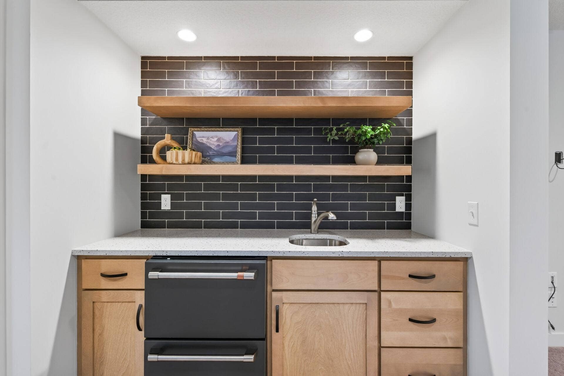 9960 Arrowwood Trail, Woodbury, MN 55129 This stylish wet bar boasts striking dark tile and floating shelves, plus a sink and fridge; the perfect spot to grab a drink after some friendly competition on the sport court.