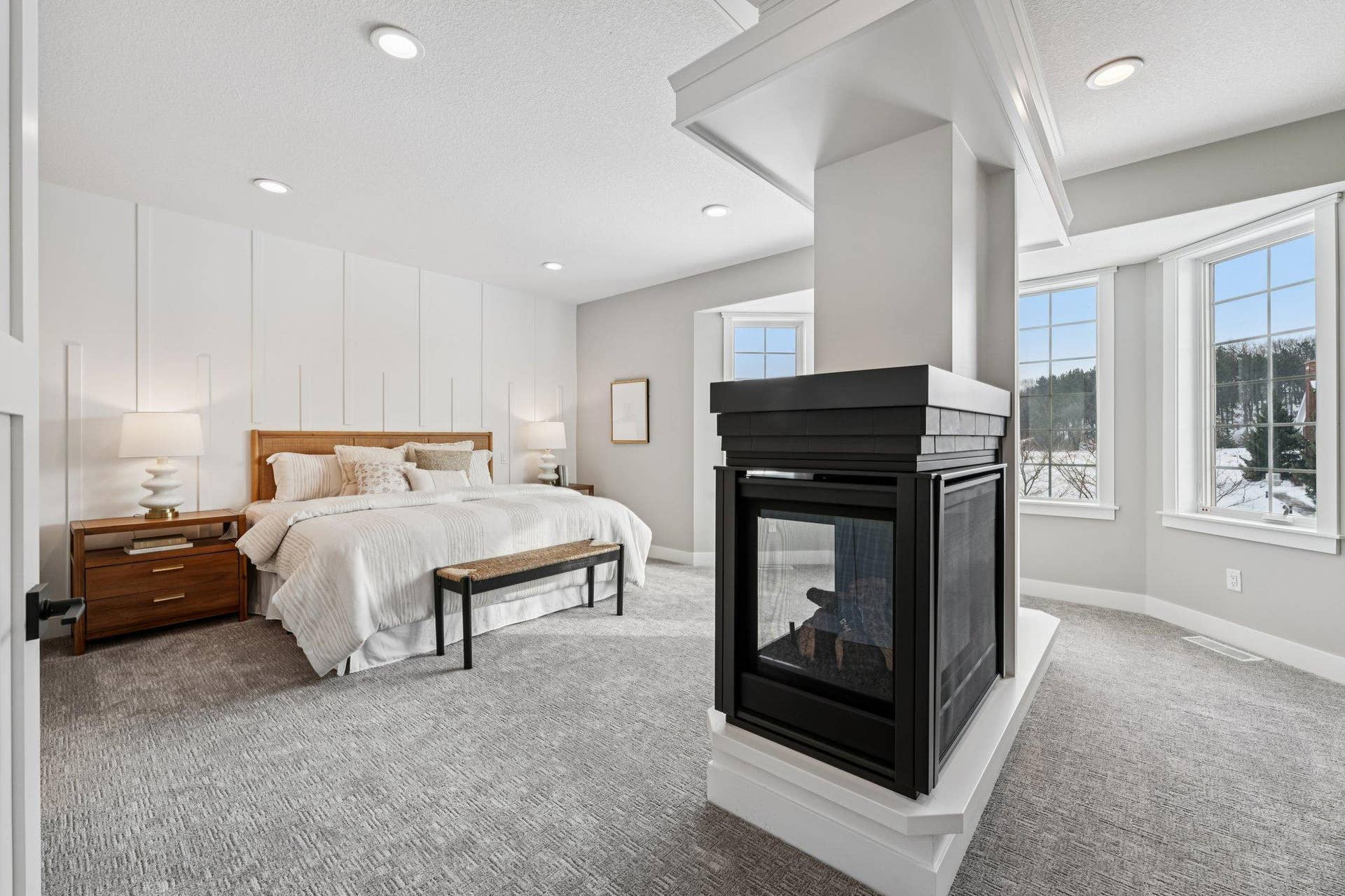 Spacious primary suite showcasing a dramatic two-sided fireplace, soft neutral tones, and oversized windows that fill the room with natural light.