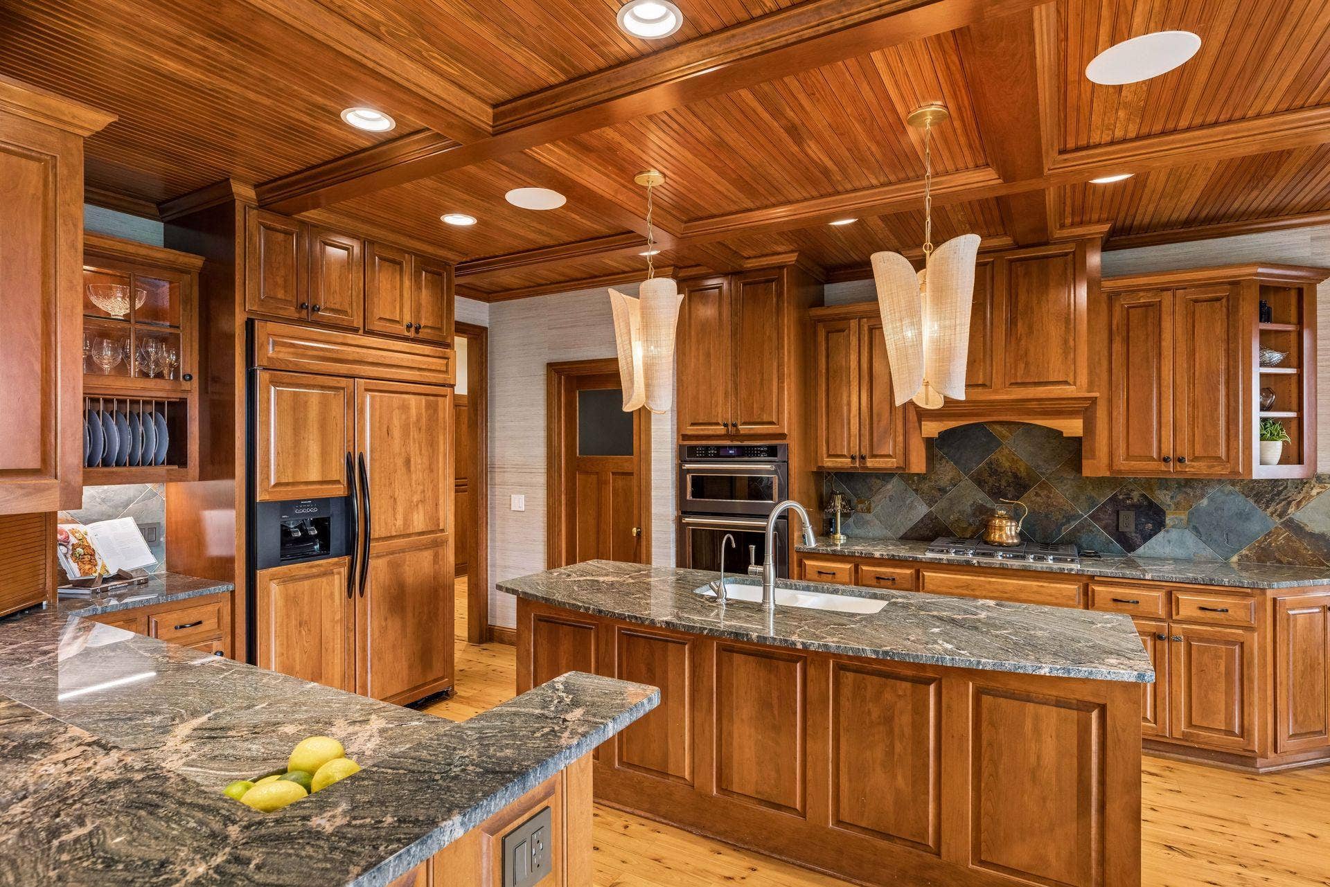 Gourmet kitchen with a coffered beadboard ceiling, custom cherry cabinetry, slate backsplash, and granite counters, opening to a cozy keeping room with a stone fireplace and porch access