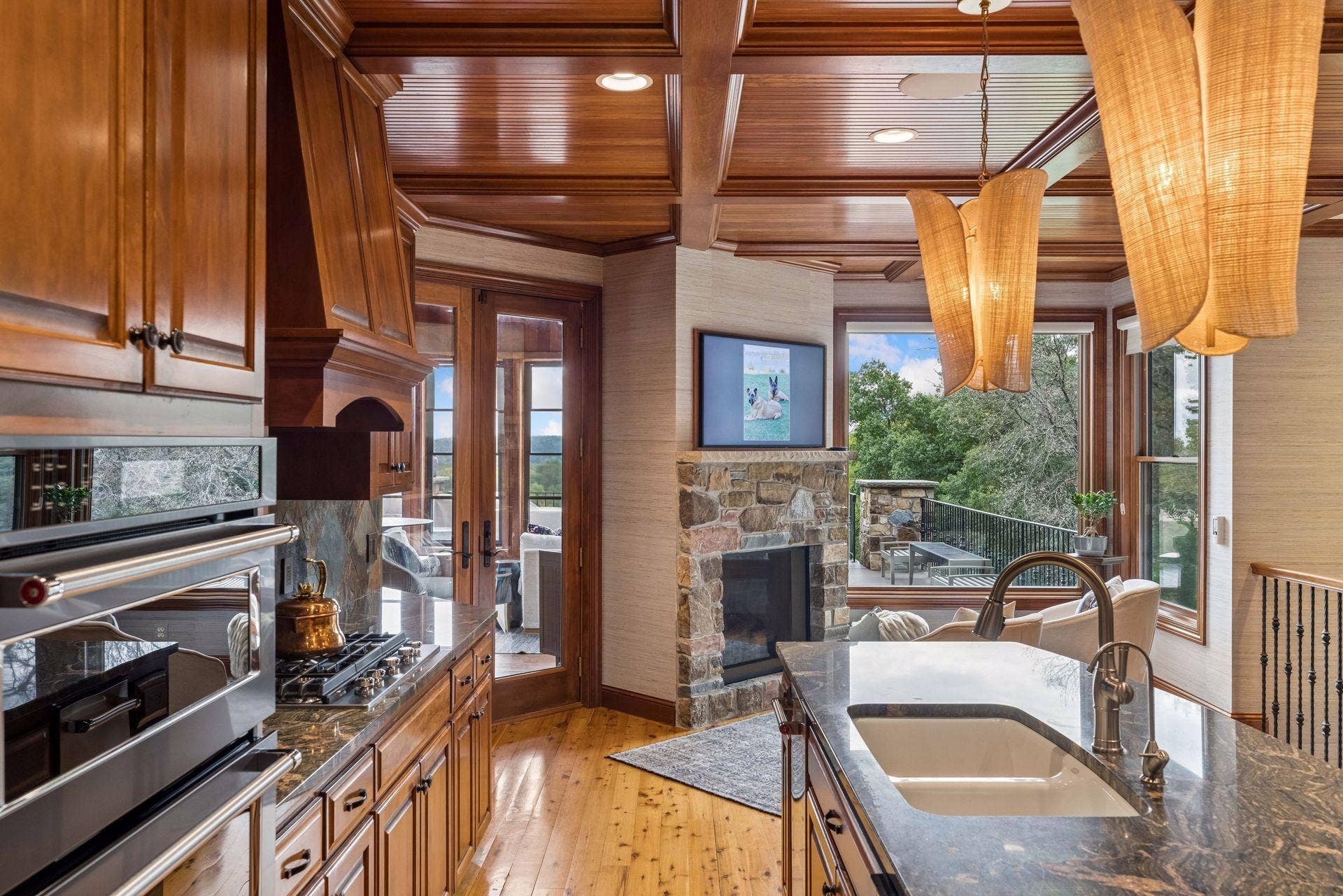 Gourmet kitchen with a coffered beadboard ceiling, custom cherry cabinetry, slate backsplash, and granite counters, opening to a cozy keeping room with a stone fireplace and porch access