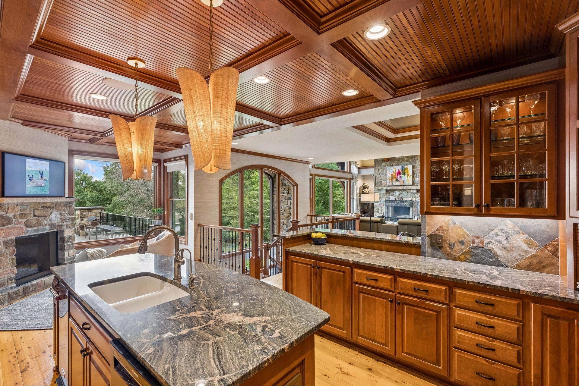 Gourmet kitchen with a coffered beadboard ceiling, custom cherry cabinetry, slate backsplash, and granite counters, opening to a cozy keeping room with a stone fireplace and porch access