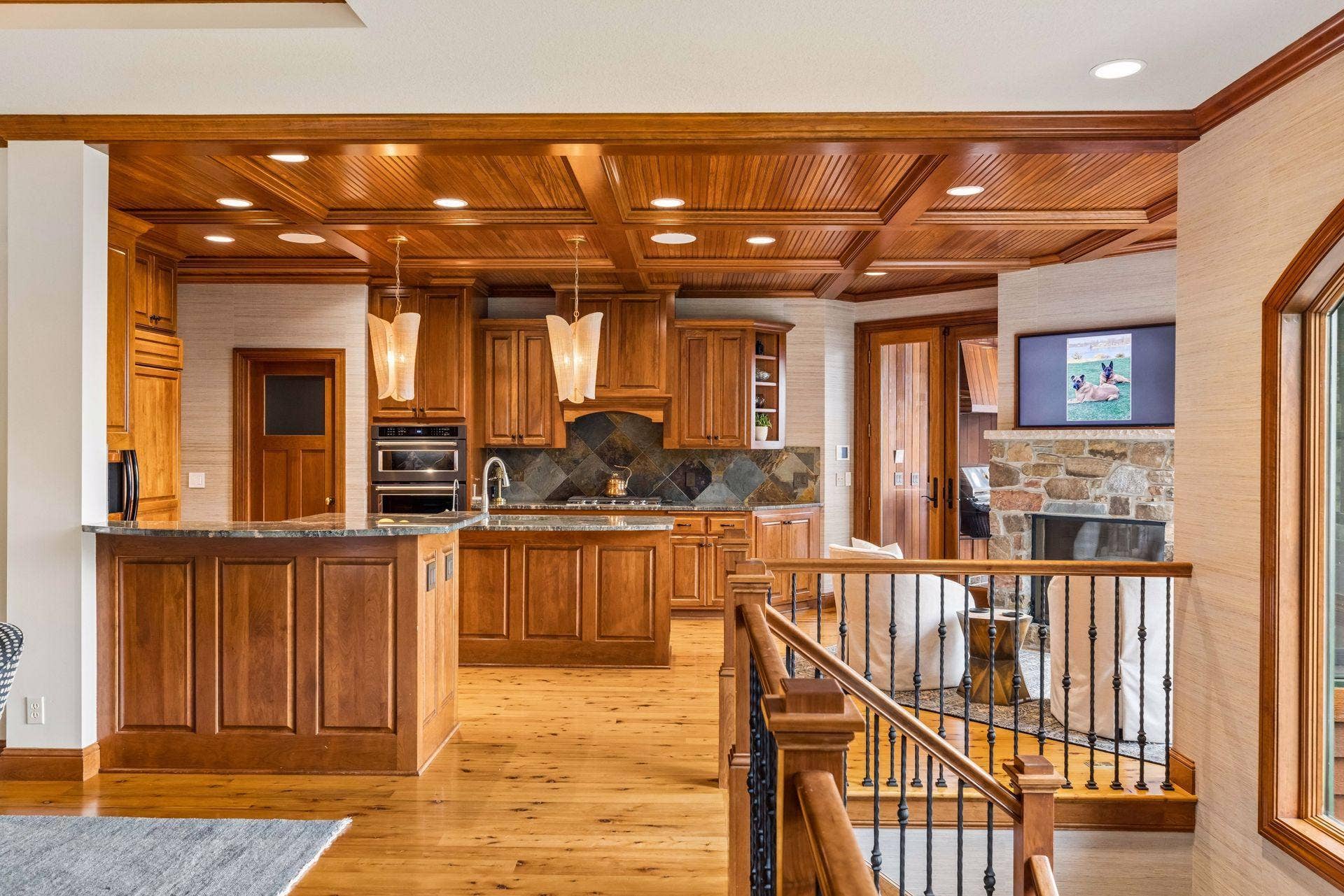 Gourmet kitchen with a coffered beadboard ceiling, custom cherry cabinetry, slate backsplash, and granite counters, opening to a cozy keeping room with a stone fireplace and porch access