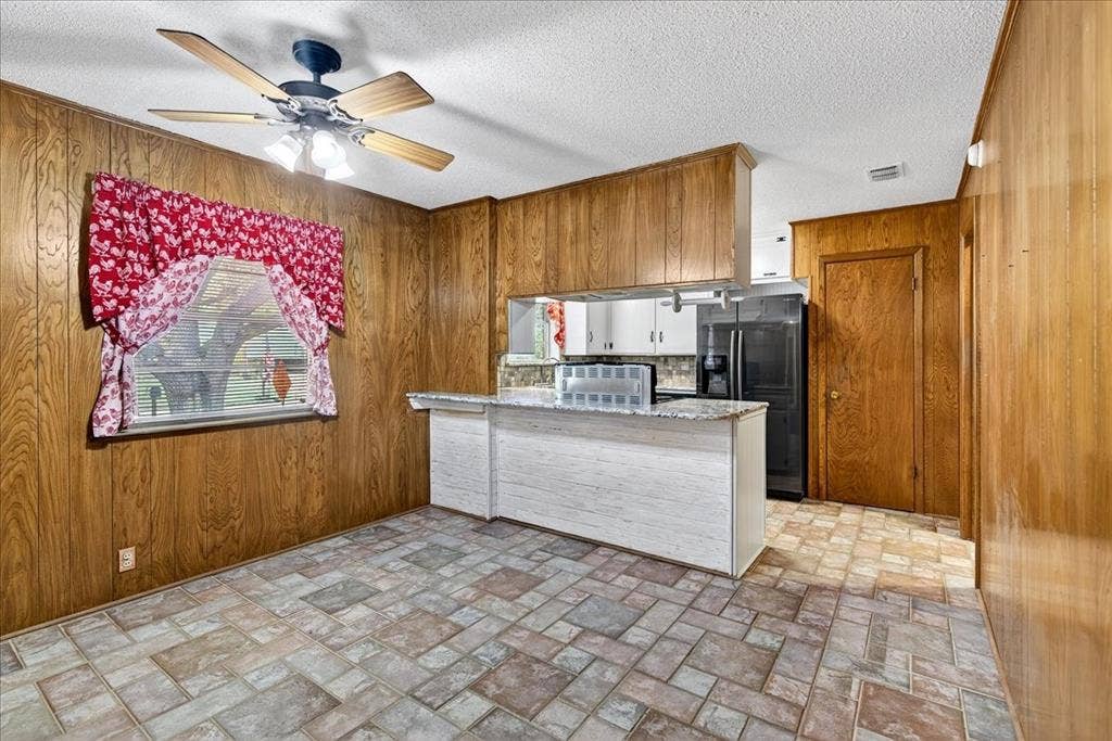 Kitchen featuring wooden walls, a peninsula, black fridge, a ceiling fan, and light stone countertops