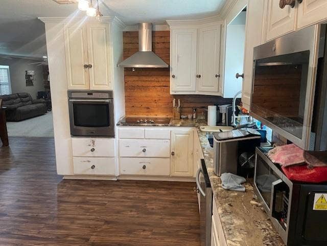 Kitchen featuring appliances with stainless steel finishes, wall chimney range hood, dark wood-type flooring, light stone countertops, and white cabinets